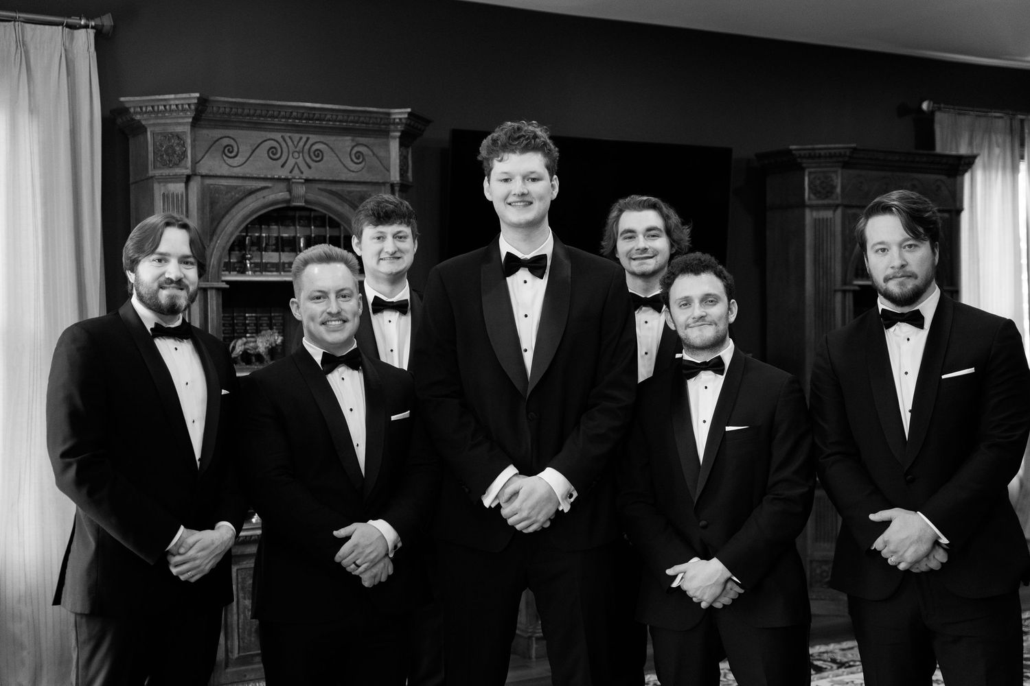 A black and white formal portrait of a wedding party in tuxedos standing together in an elegant indoor setting.