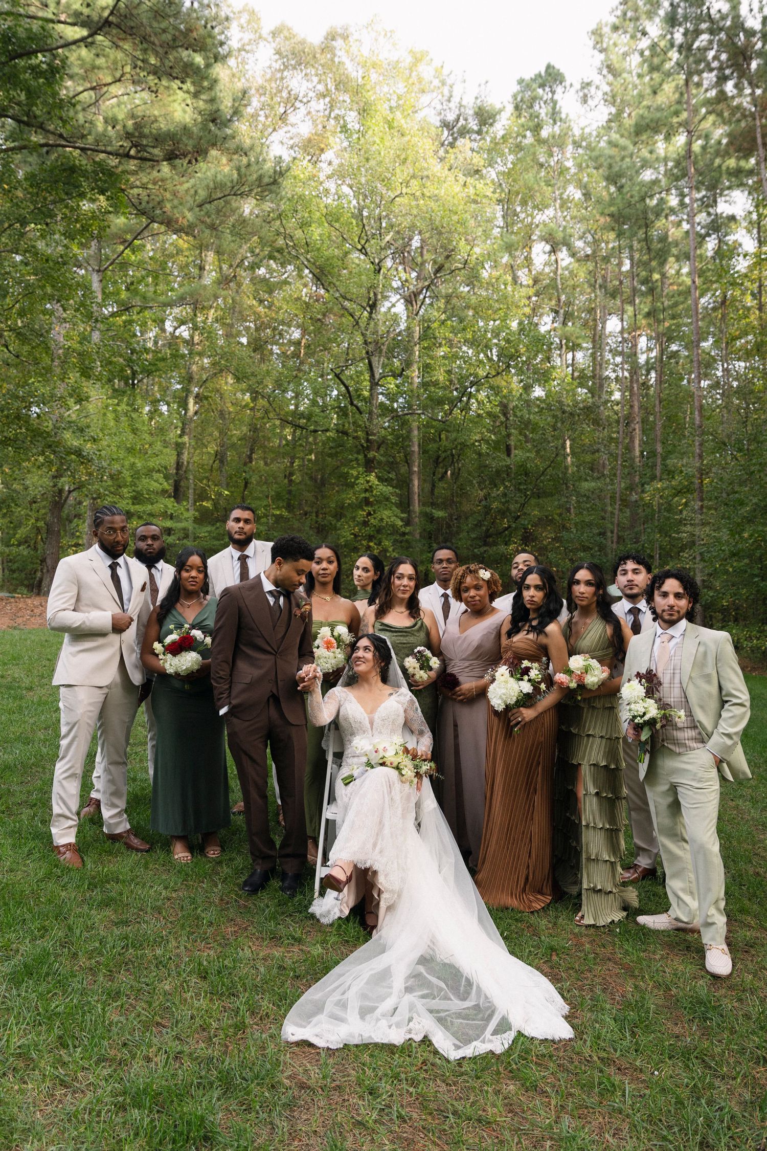 Wedding party poses outdoors in earth-toned attire with white and brown color scheme against a wooded backdrop.