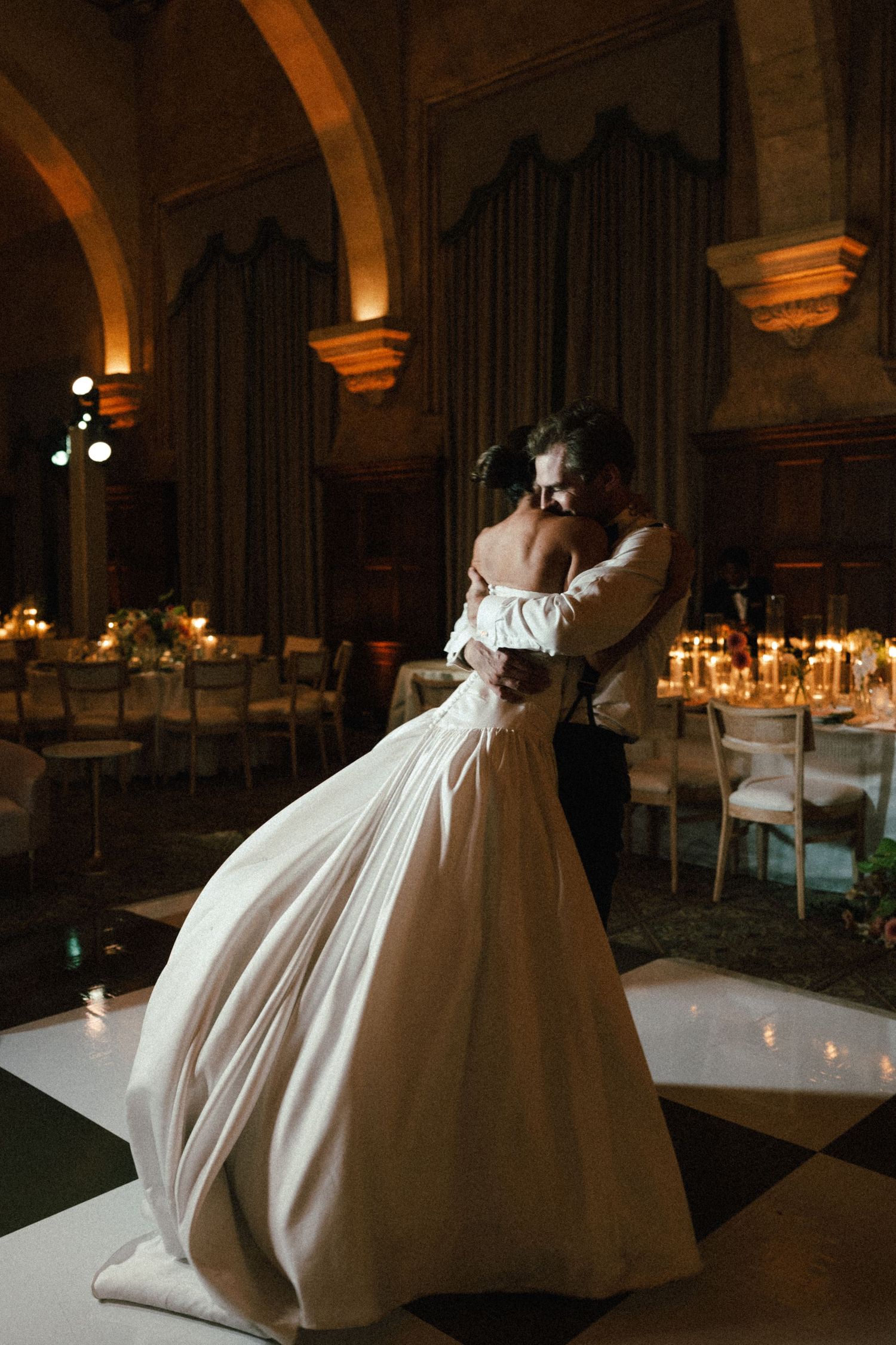Bride and groom share romantic first dance in dimly lit elegant ballroom with candlelit reception tables.
