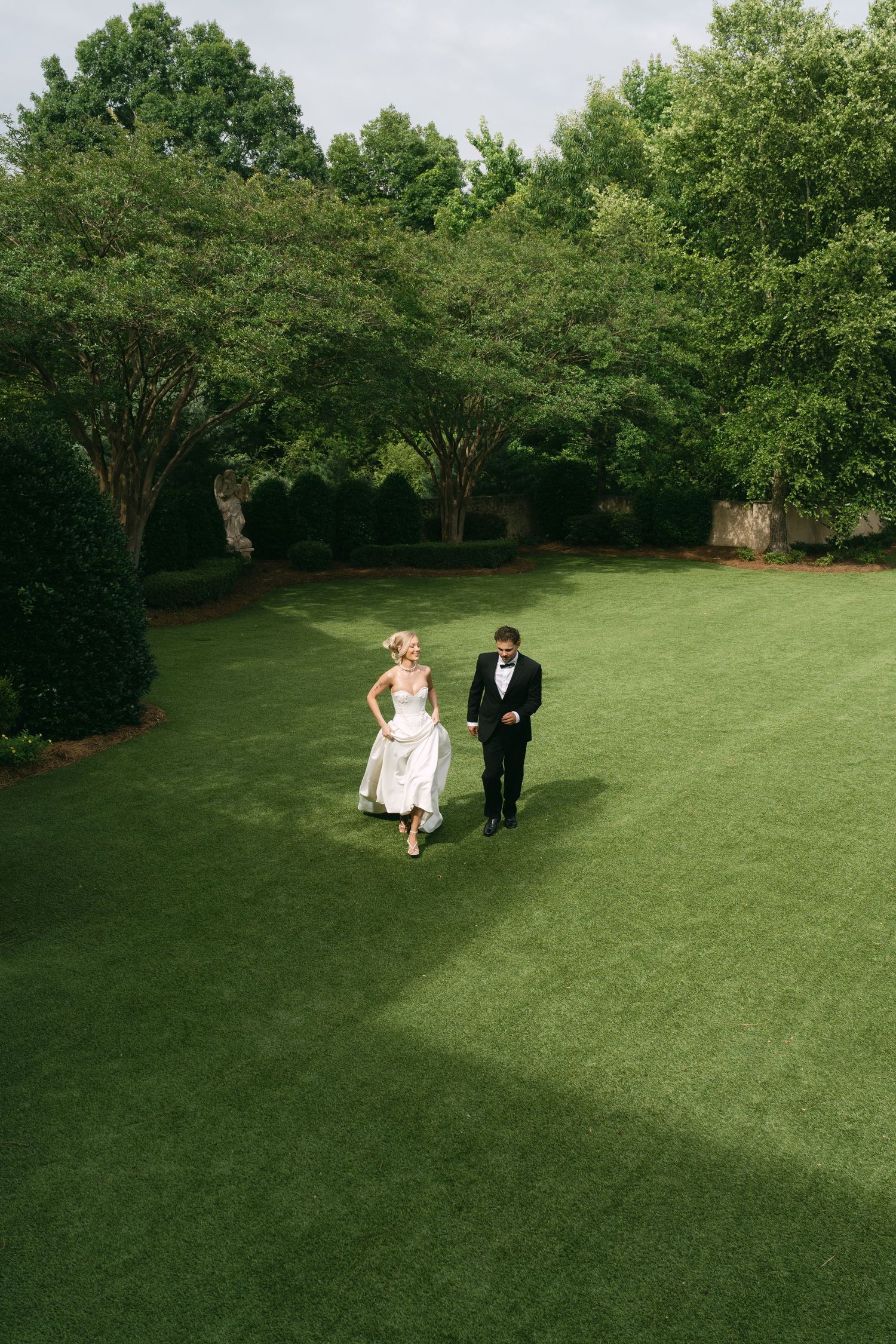 Bride and groom walking hand in hand across lush green lawn surrounded by trees on their wedding day.