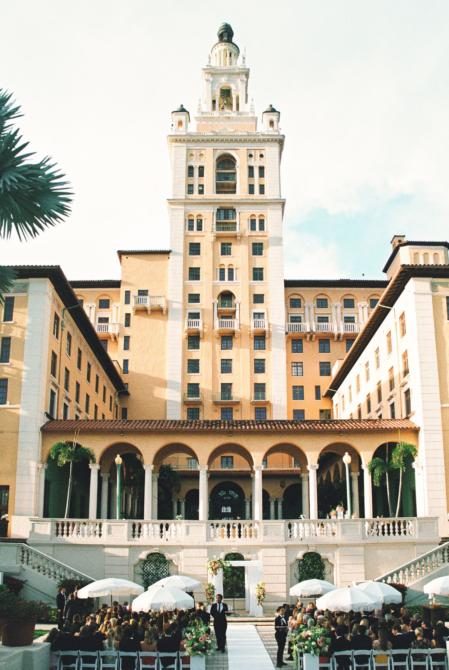 Grand historic hotel tower serves as backdrop for outdoor wedding ceremony with white umbrellas and guests.