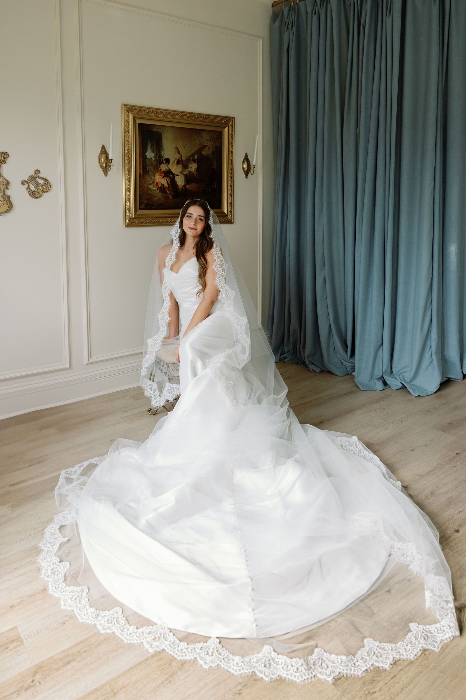 Elegant bride in white lace wedding gown with long train and veil poses in front of teal curtains and floral wall art.