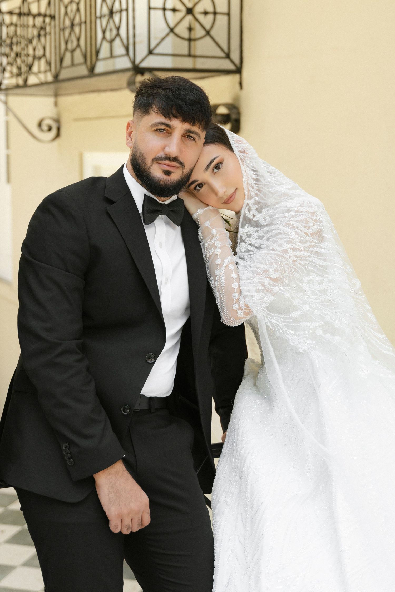 A romantic wedding couple poses together in formal attire against a light-colored architectural backdrop.
