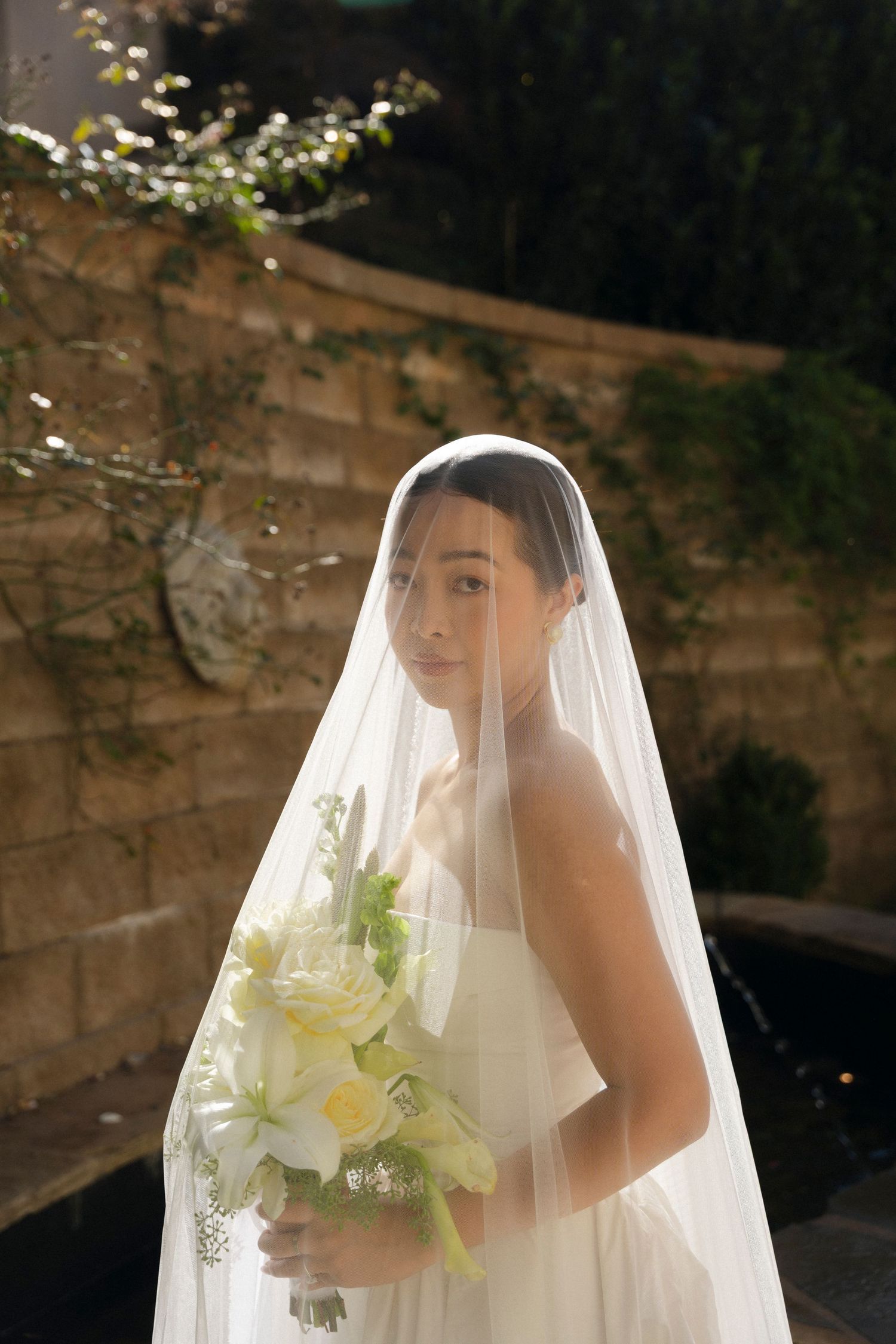 A bride in a white veil holds a delicate bouquet of white lilies against a dark background.