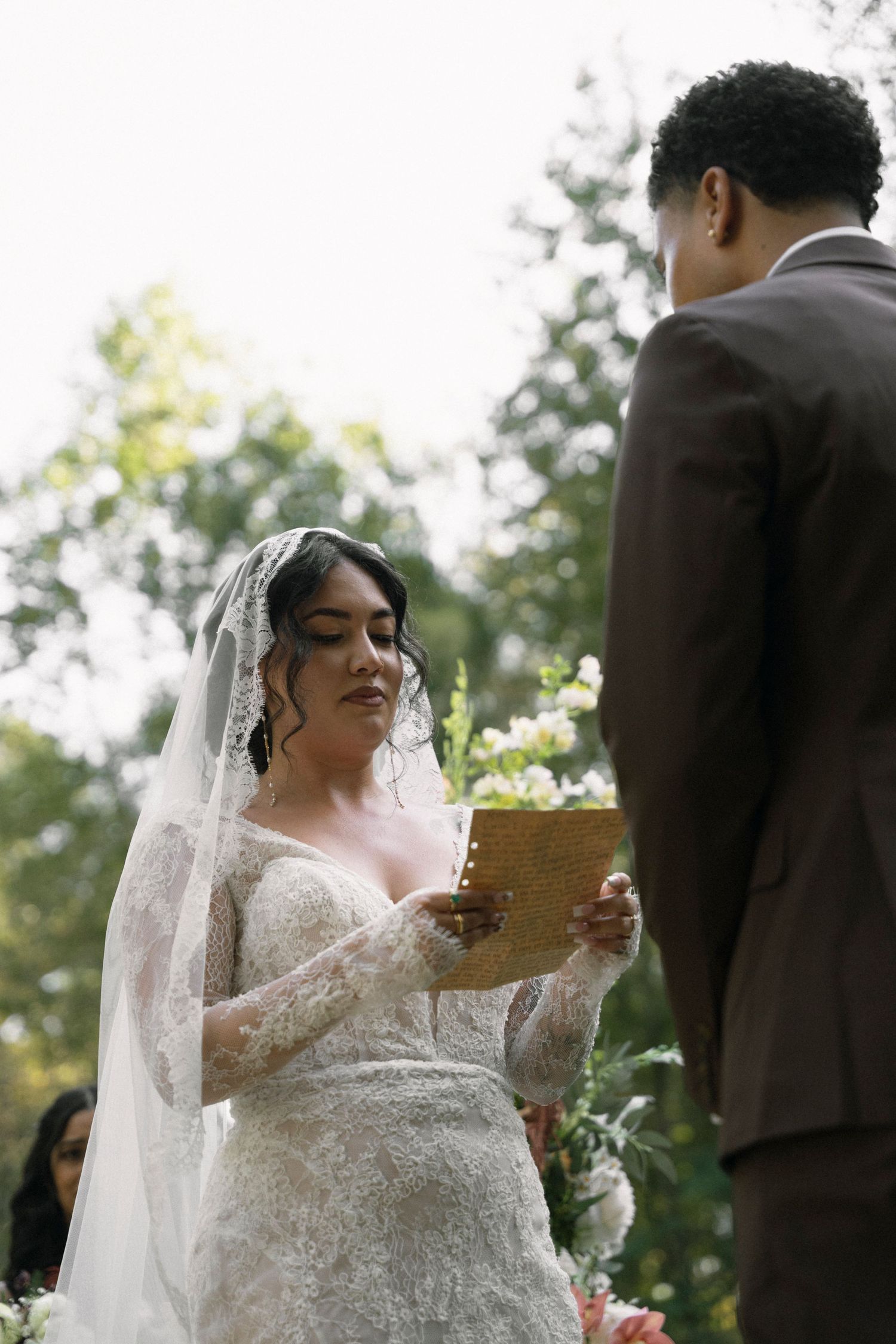 Bride in elegant white lace gown and veil reads from a paper during an outdoor wedding ceremony.
