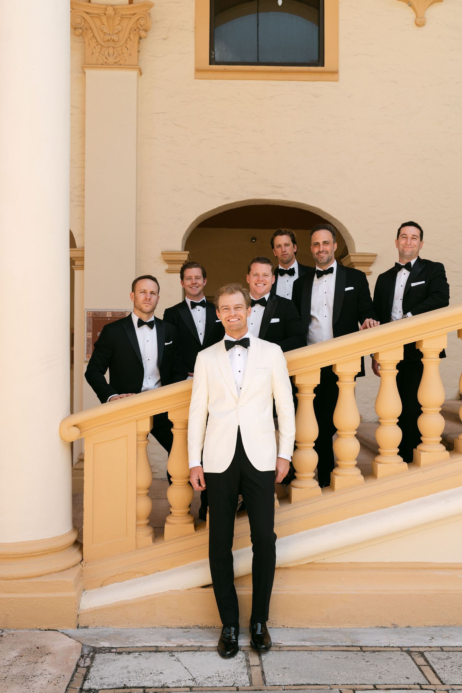 Groom in white tuxedo jacket stands before groomsmen in black tuxedos on elegant staircase at wedding venue.