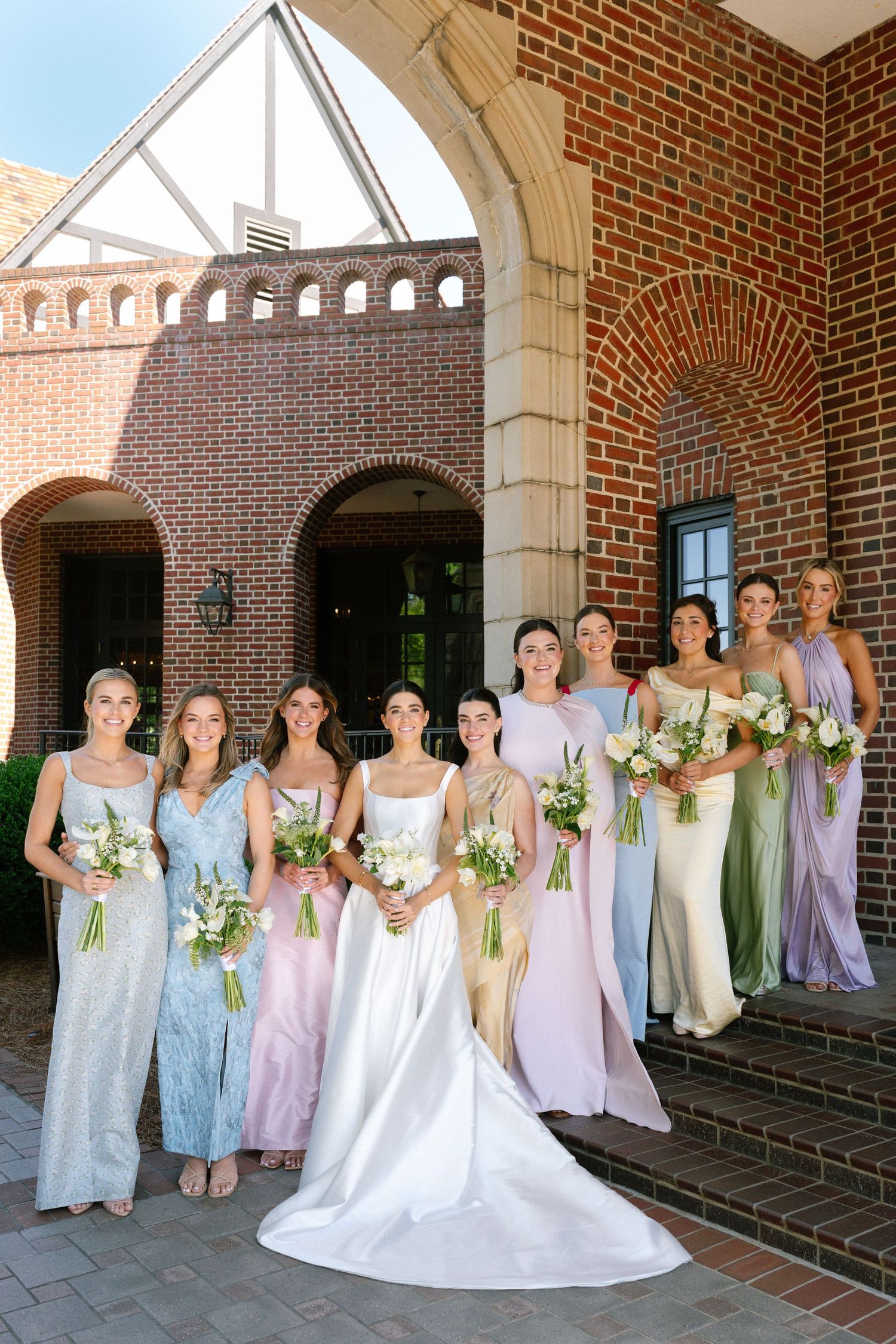 Bride and bridesmaids in colorful pastel gowns holding bouquets outside a brick church.