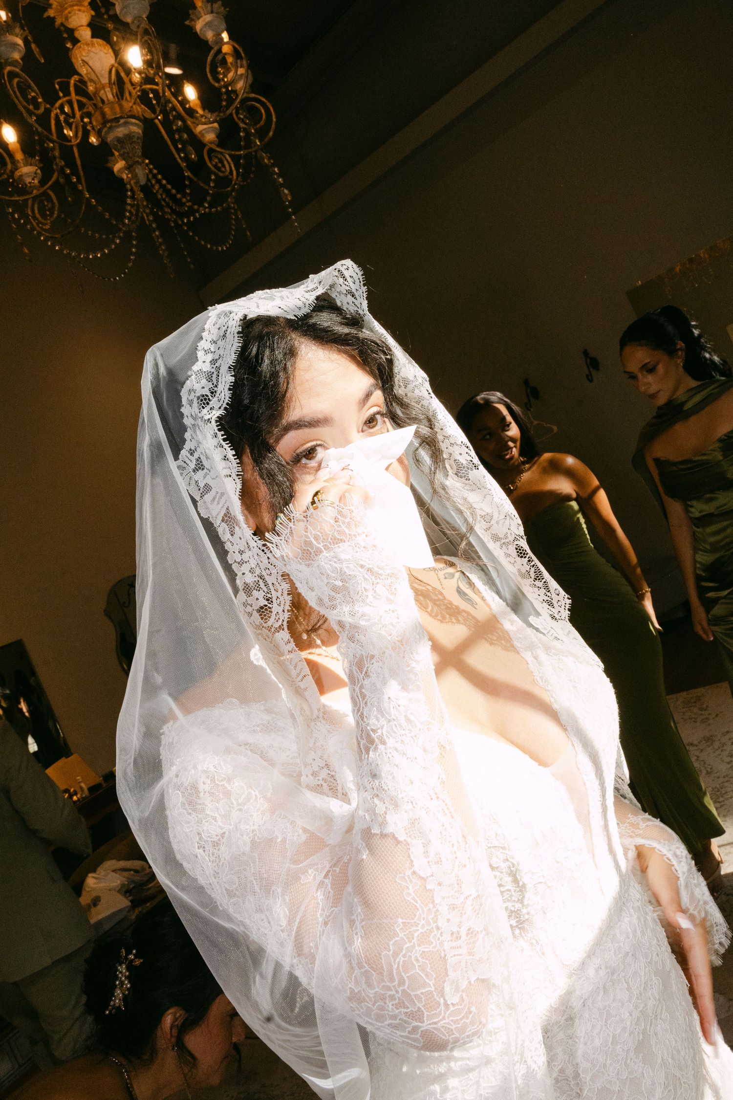 A bride in an elegant white lace wedding dress and veil poses dramatically against a dark background.
