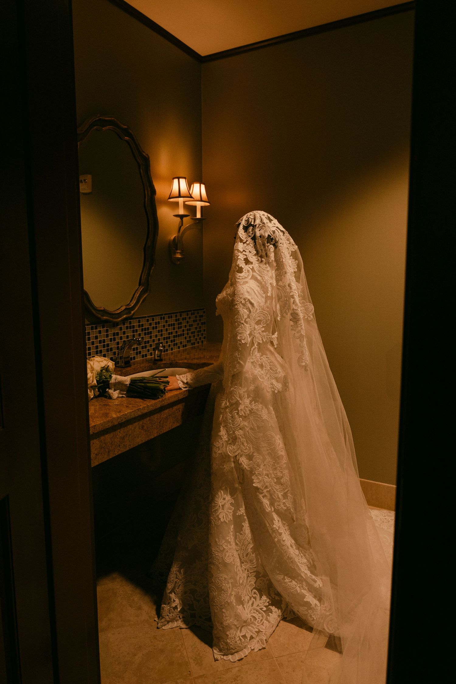 A white lace wedding dress hangs elegantly in a dimly lit room with warm lighting and dark walls.