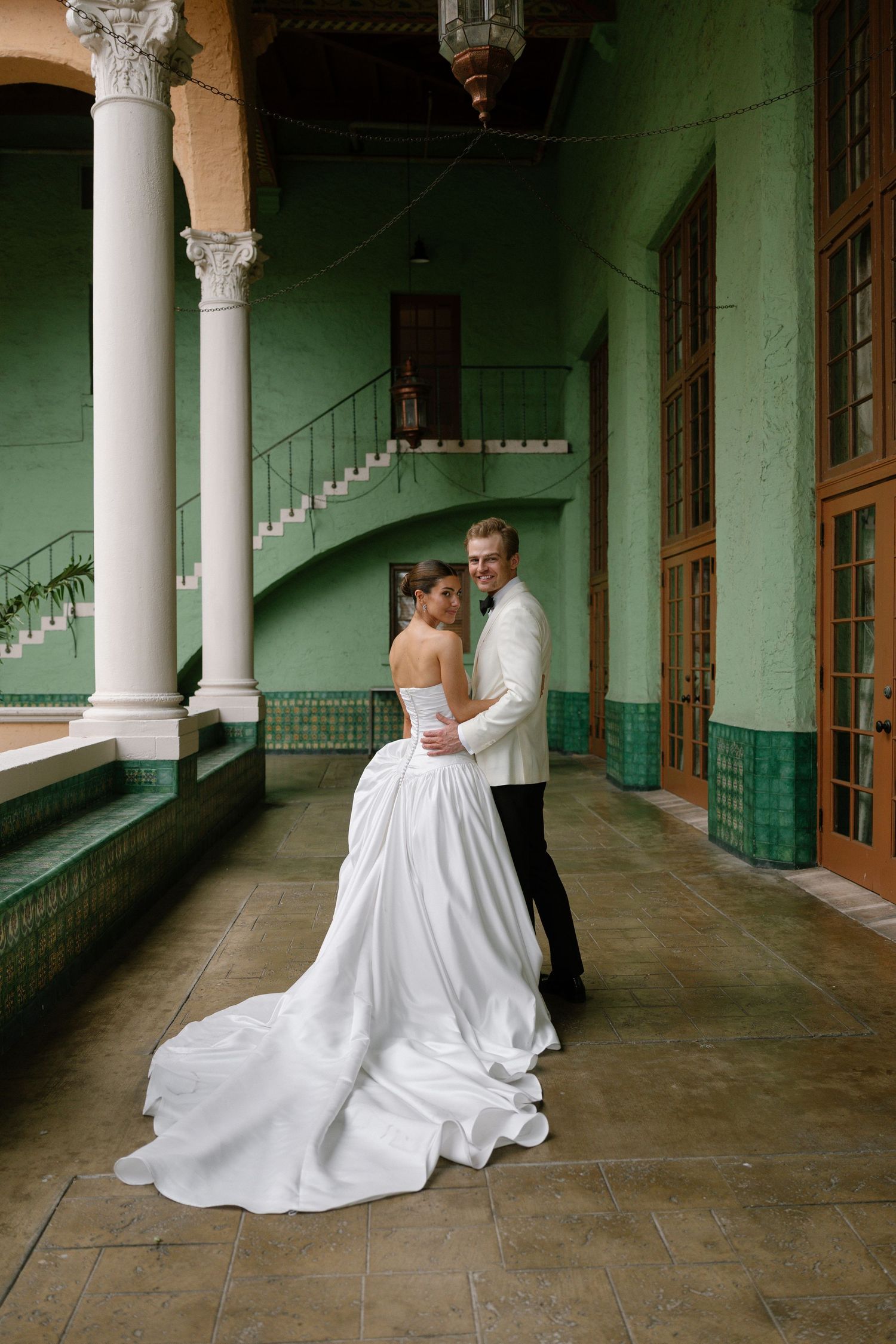 Bride in strapless white gown with long train embraces groom in white jacket in green-walled courtyard.