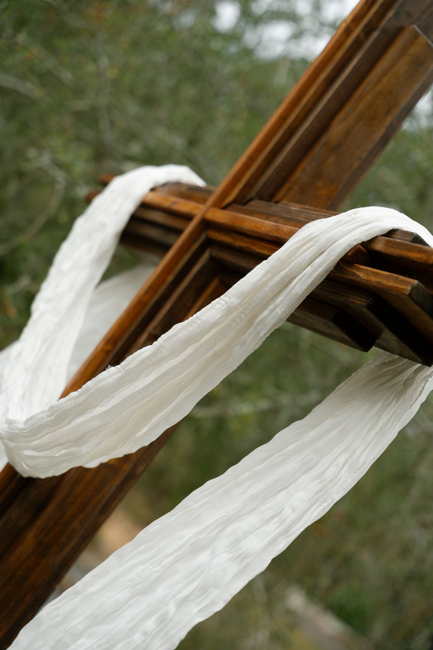 A wooden cross draped with flowing white fabric against a blurred green outdoor background.