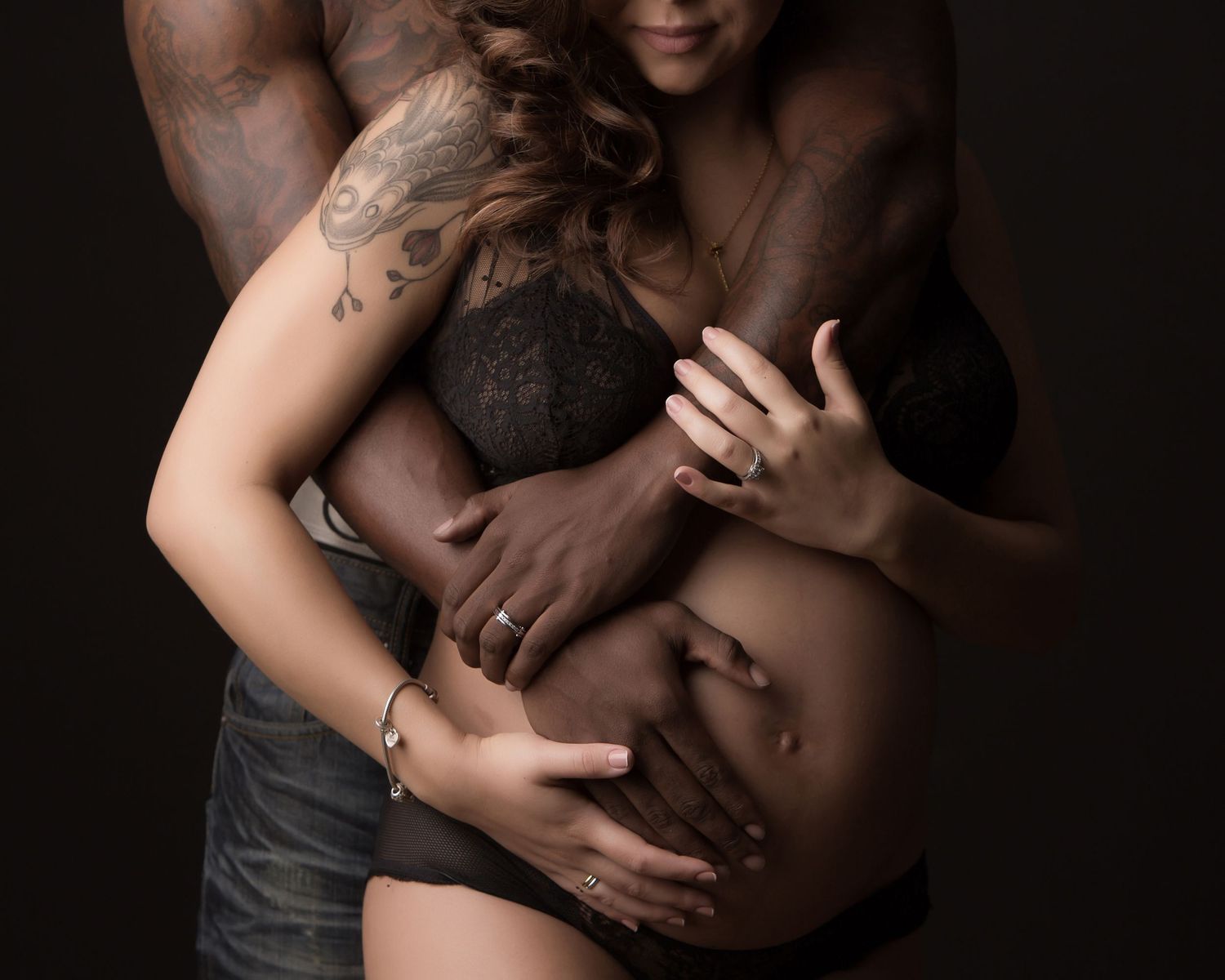 Intimate artistic photograph of an expectant mother being embraced from behind, showcasing a maternity portrait against dark backdrop.