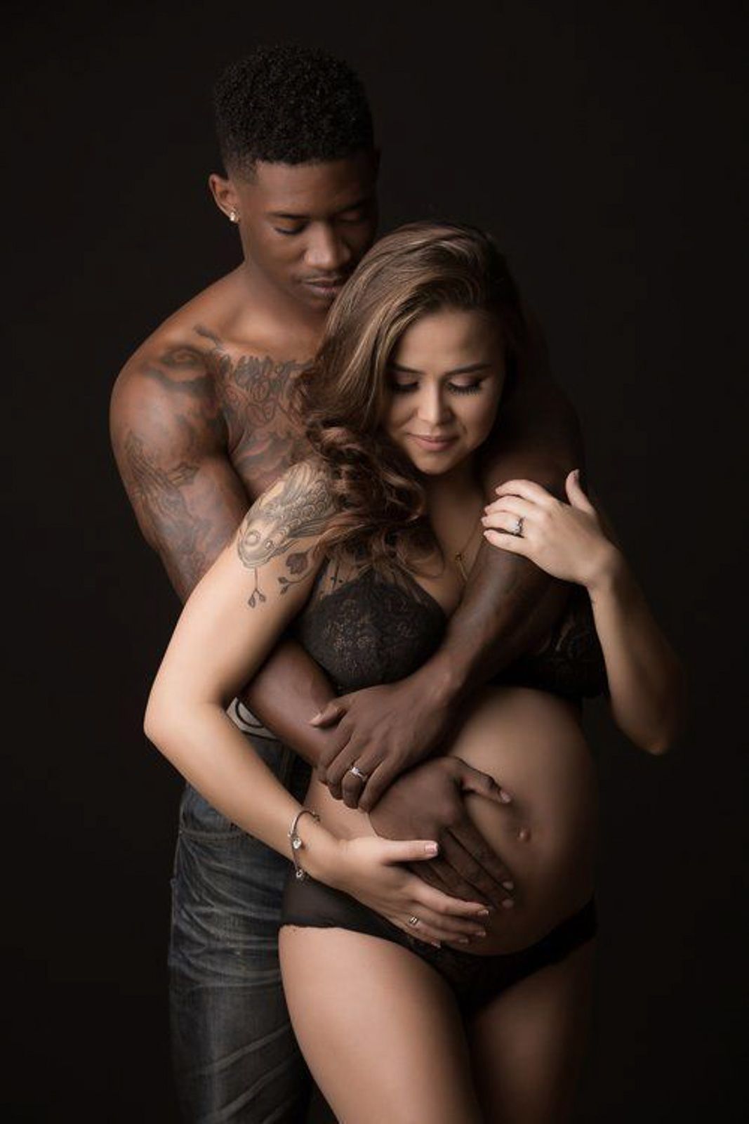Couple sharing a tender maternity photo, man embracing tattooed pregnant woman against dark background.
