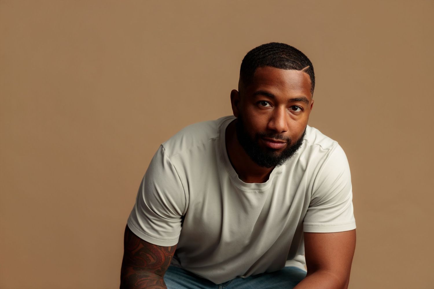 A young Black man in a white t-shirt poses confidently against a warm tan background, showcasing a casual yet stylish look.