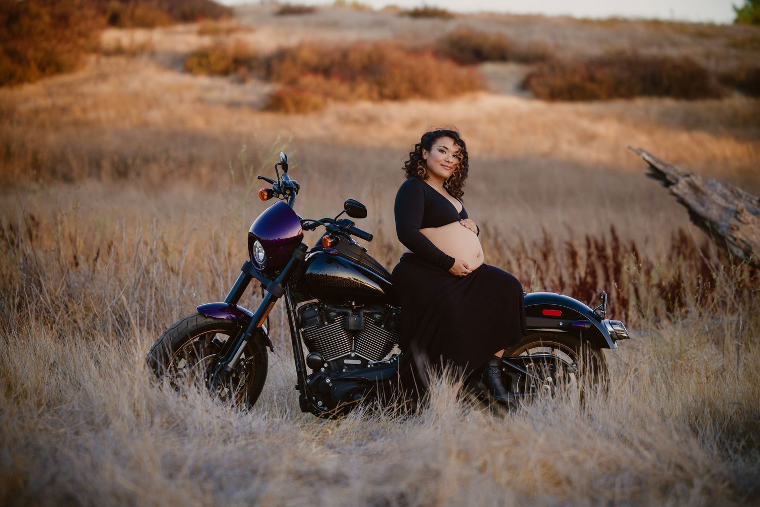 Pregnant woman in black dress poses on purple motorcycle in golden field during maternity photo shoot.