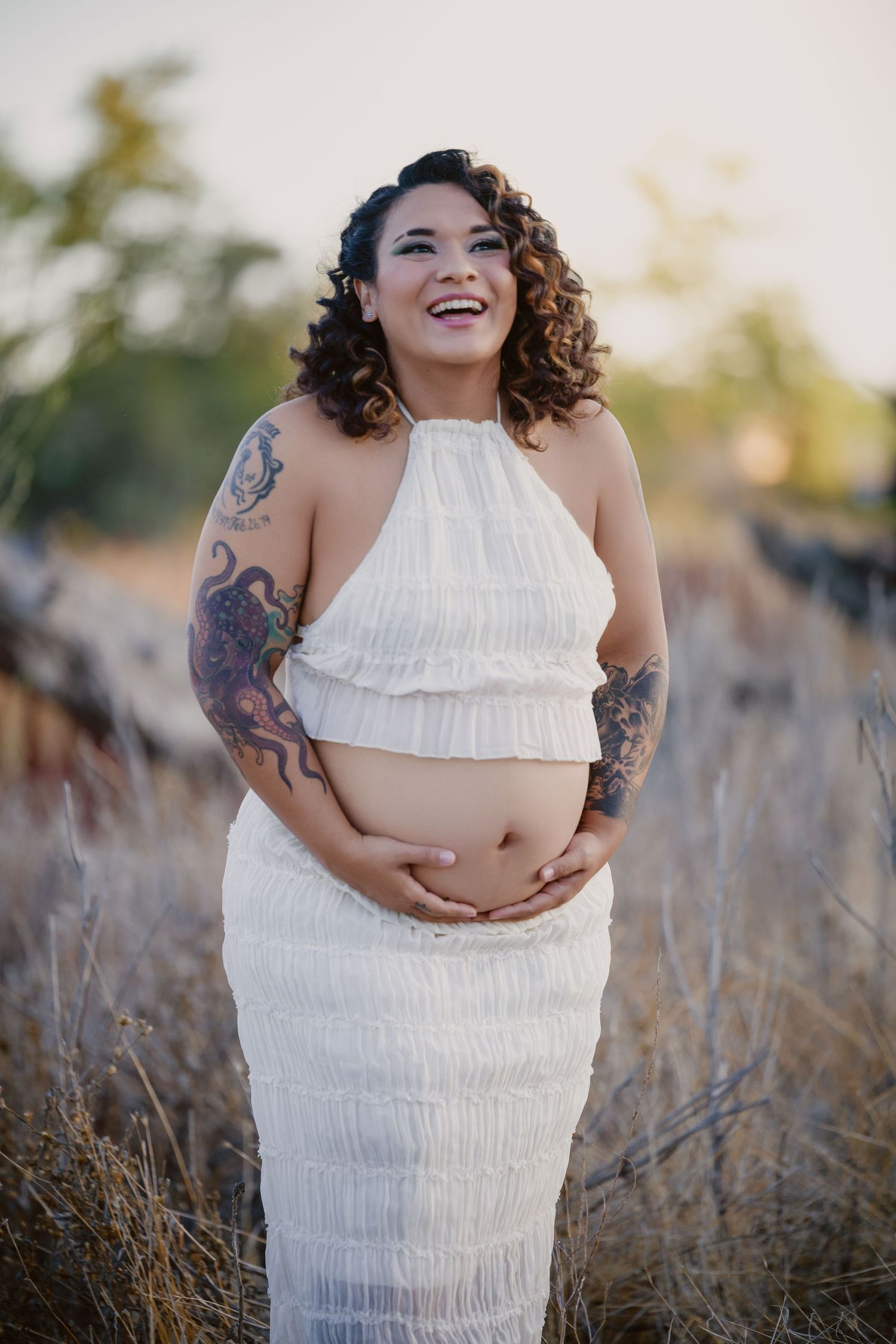 Smiling pregnant woman with tattoos in white crop top and skirt holding baby bump outdoors in golden field.