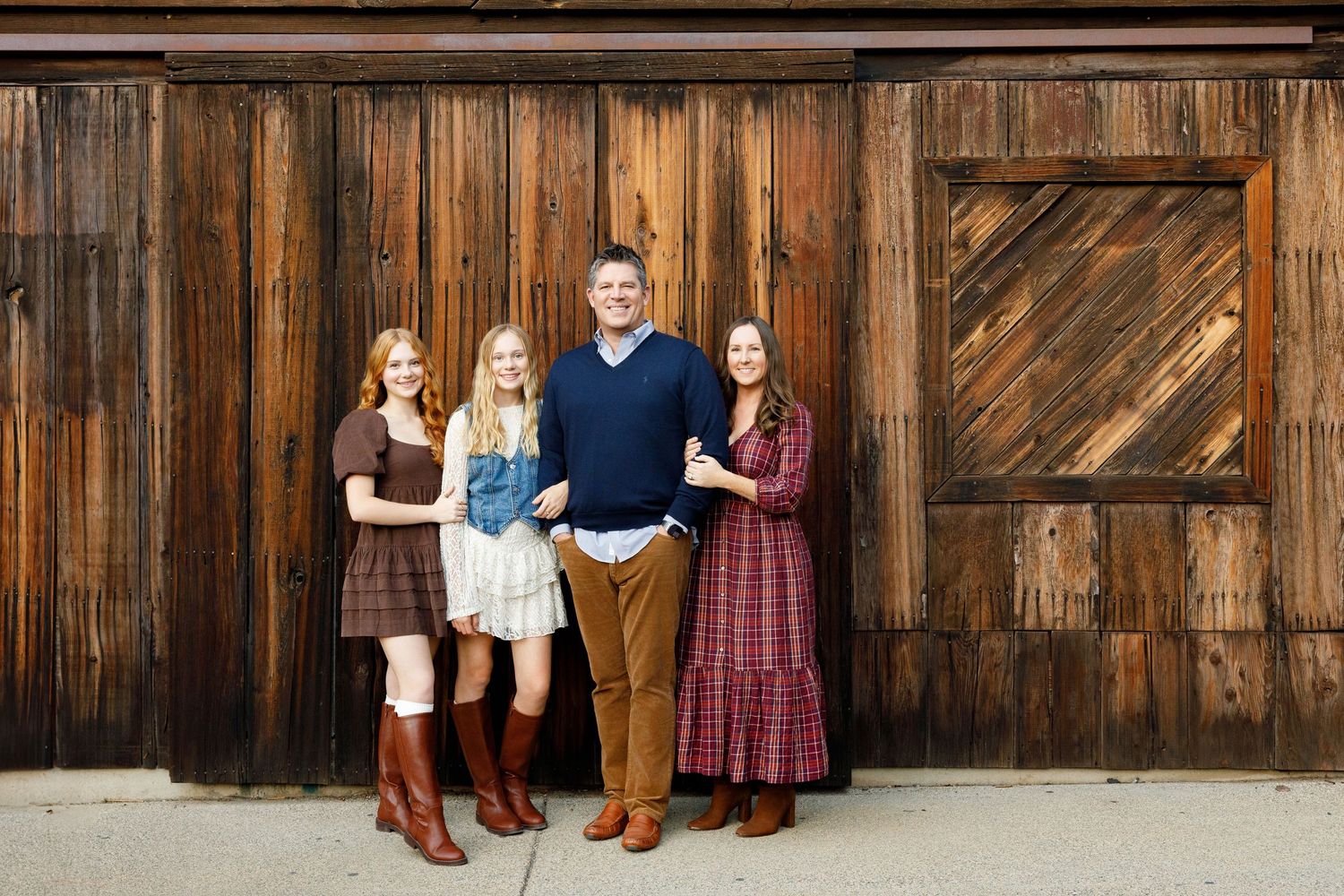 Family members in coordinating outfits pose against rustic wooden barn wall for outdoor portrait session.