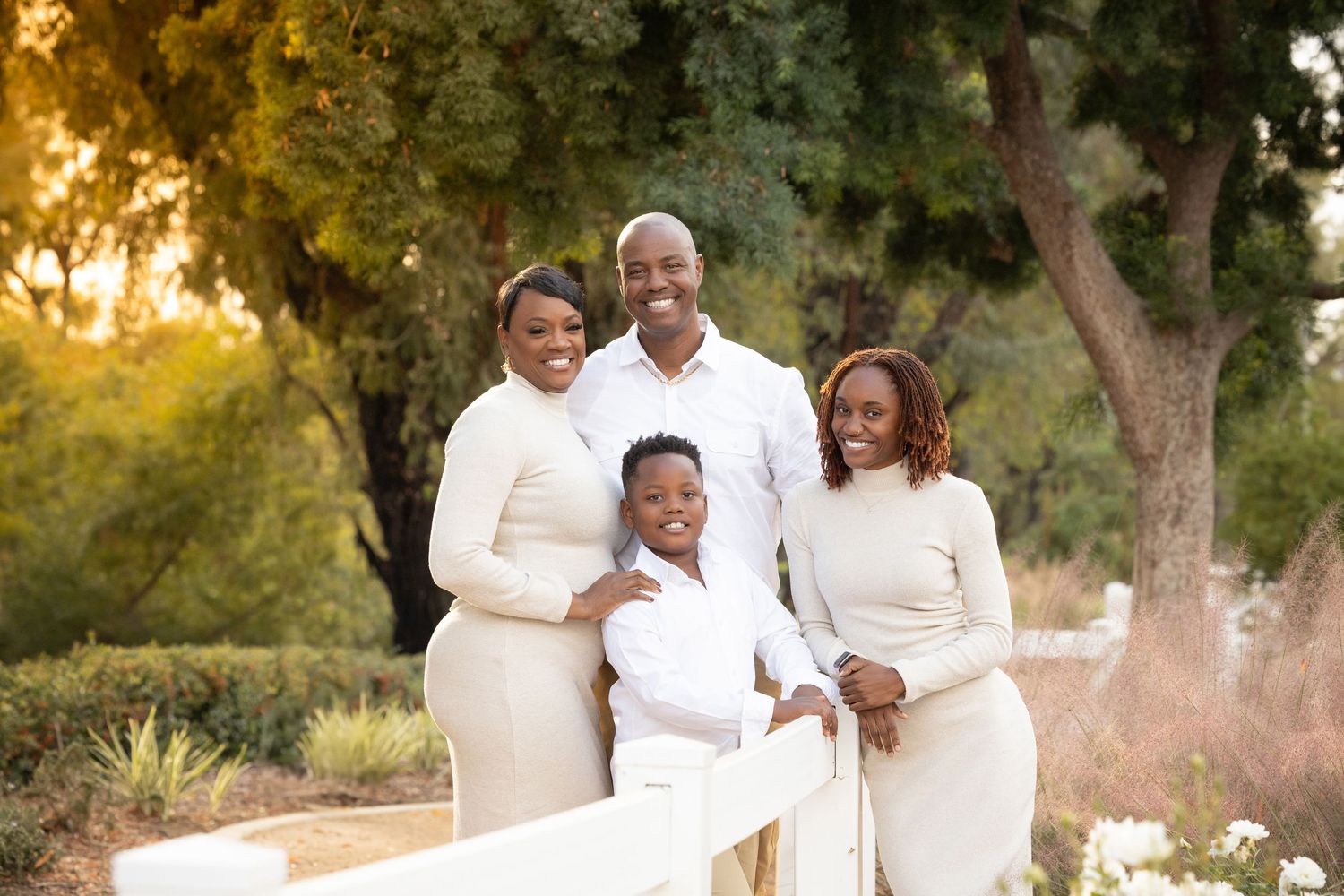 Happy Black family of four posing outdoors in matching cream outfits during golden hour in a lush garden.