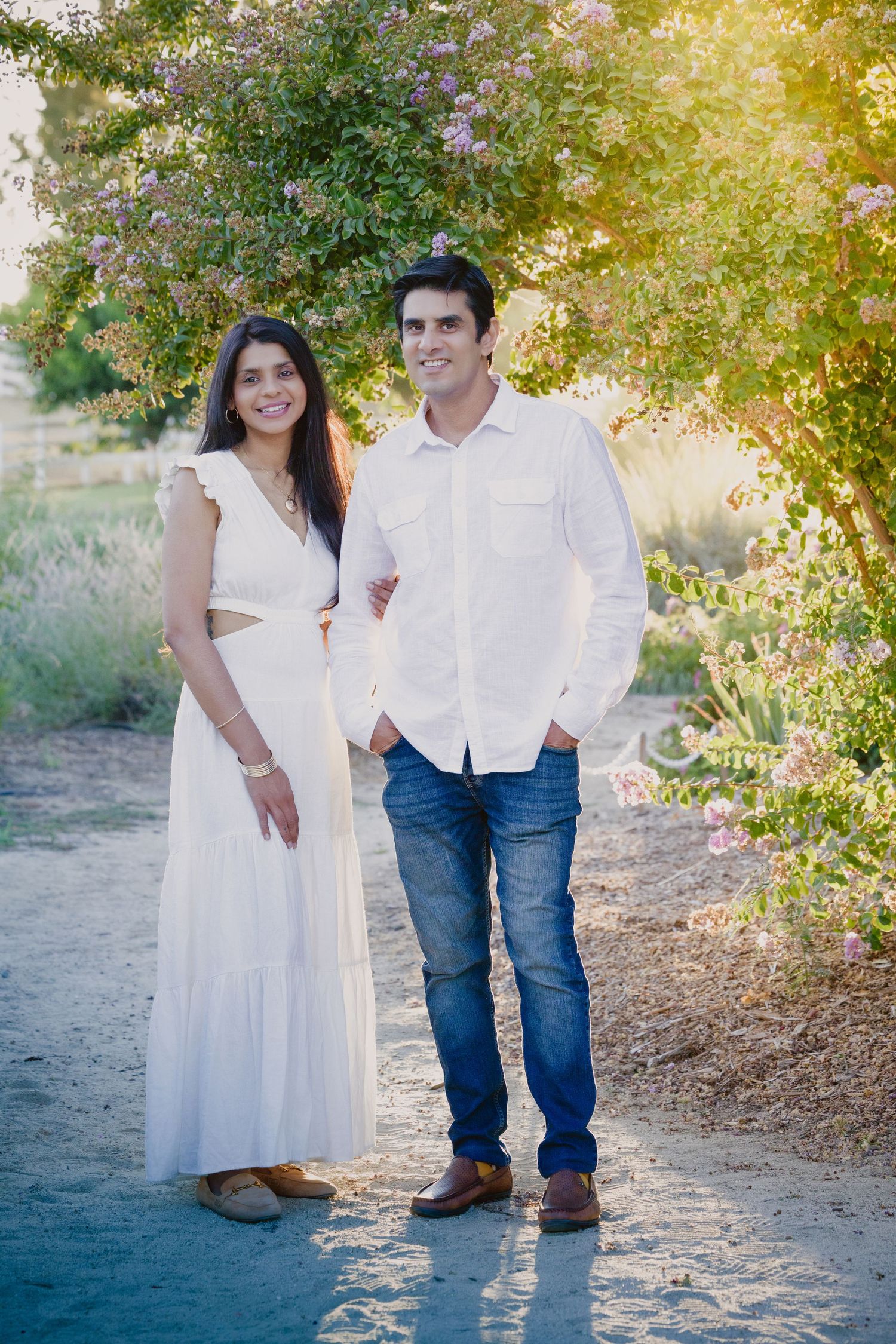 A couple stands together on a dirt path with flowering trees and golden sunlight in the background.