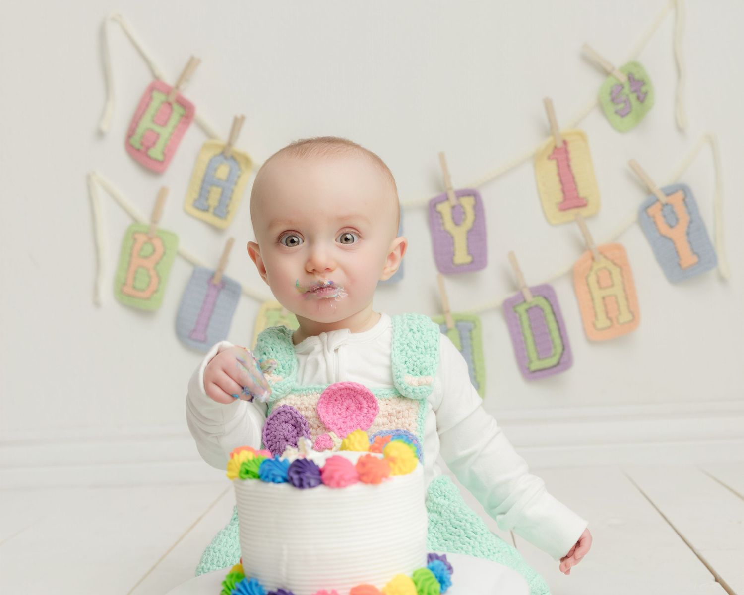 Baby girl in mint dress celebrates first birthday with colorful cake and happy birthday banner.