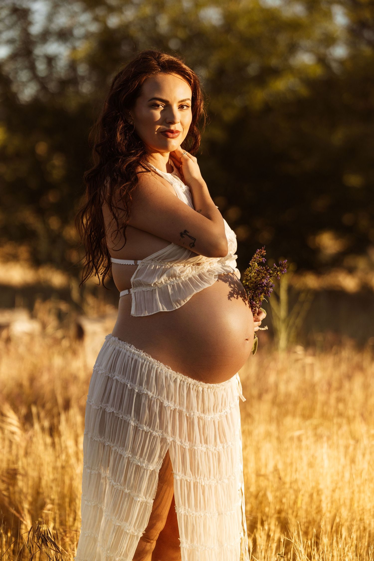Pregnant woman in white dress holding wildflowers in golden field at sunset.