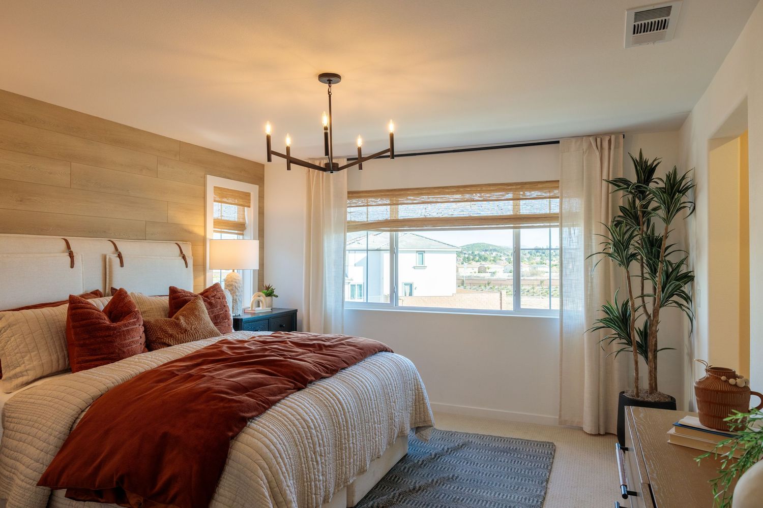 Modern bedroom with rust colored bedding, wood accent wall, black chandelier and large window with natural light.