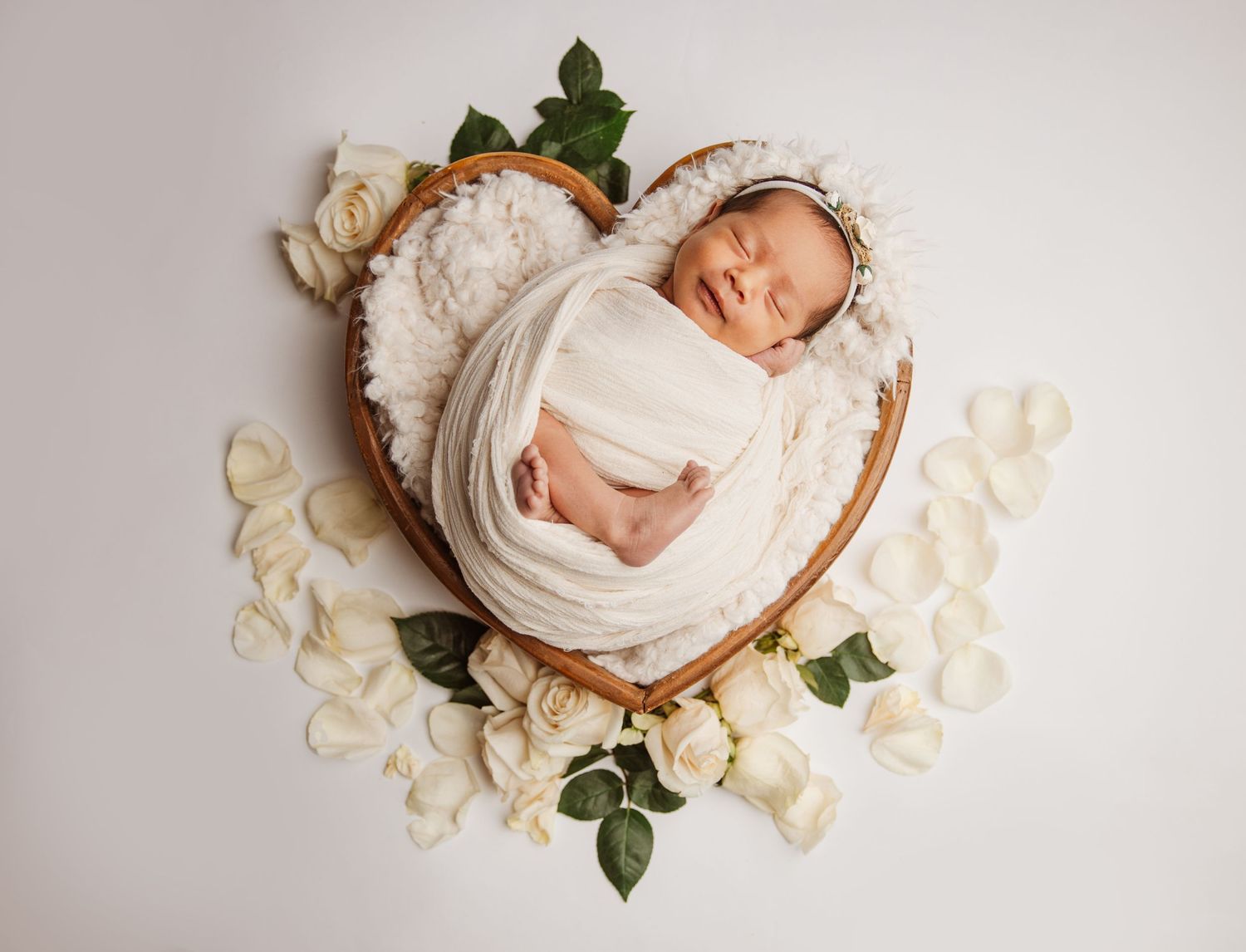 A series of newborn photography images showing a baby in soft white wraps surrounded by delicate rose petals and greenery.