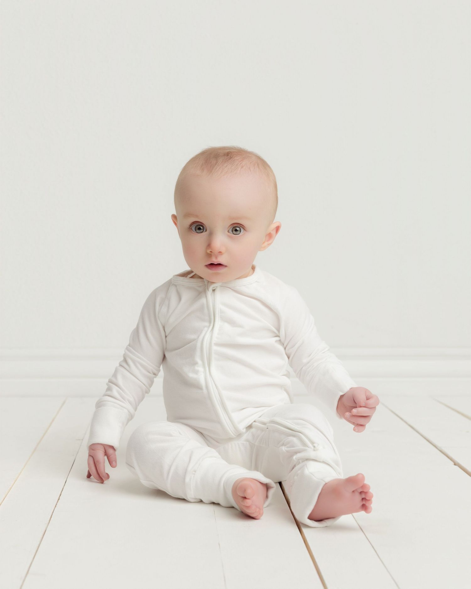 Adorable baby in white zip-up romper sits on white wooden floor against clean background.