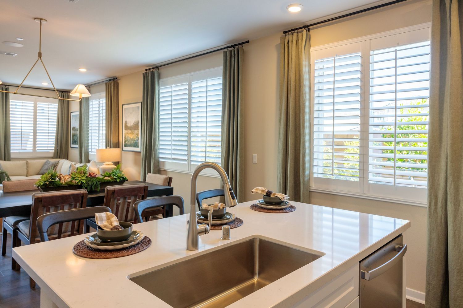 Modern kitchen with white quartz countertop, stainless sink, and plantation shutters overlooking dining area.