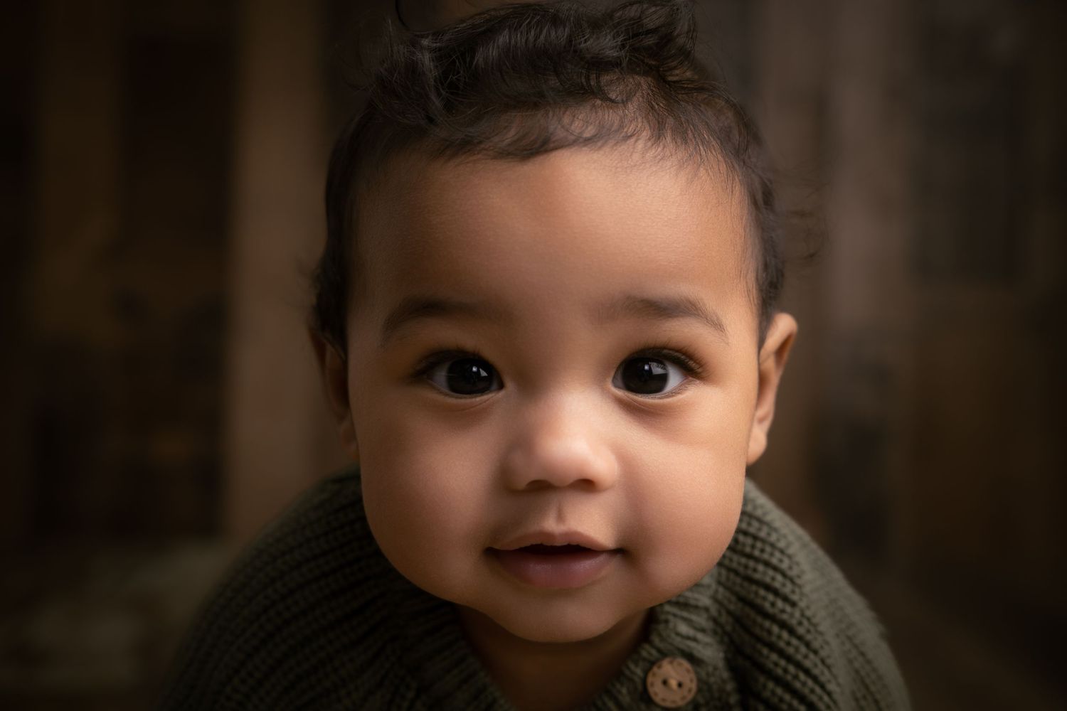 Adorable baby with big brown eyes wearing a green knit sweater, posed against a dark rustic background.