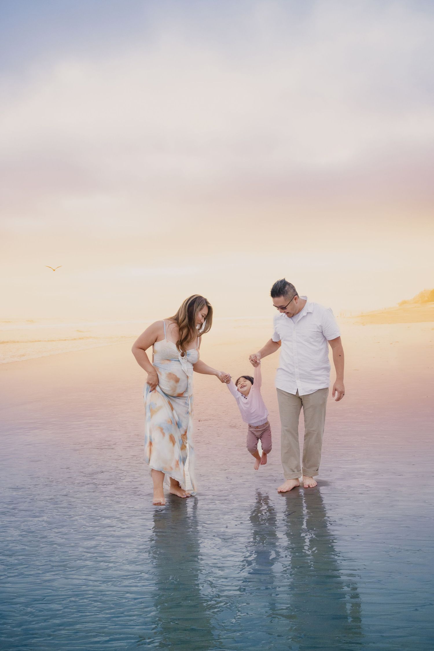 Family of three playing at the beach during sunset, parents holding baby's hands over shallow water.