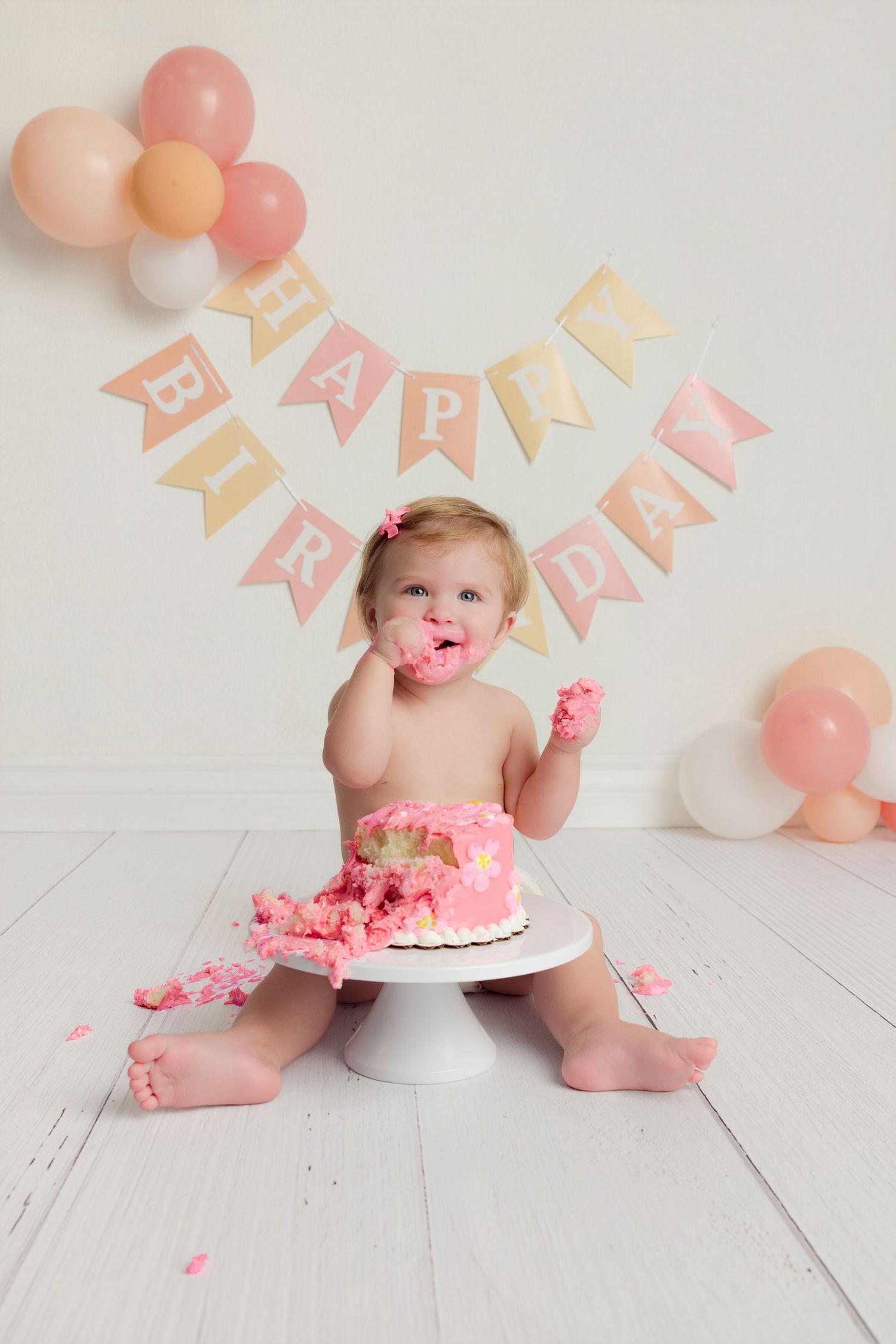 Baby girl smashing pink first birthday cake with Happy Birthday banner and balloons in white studio.