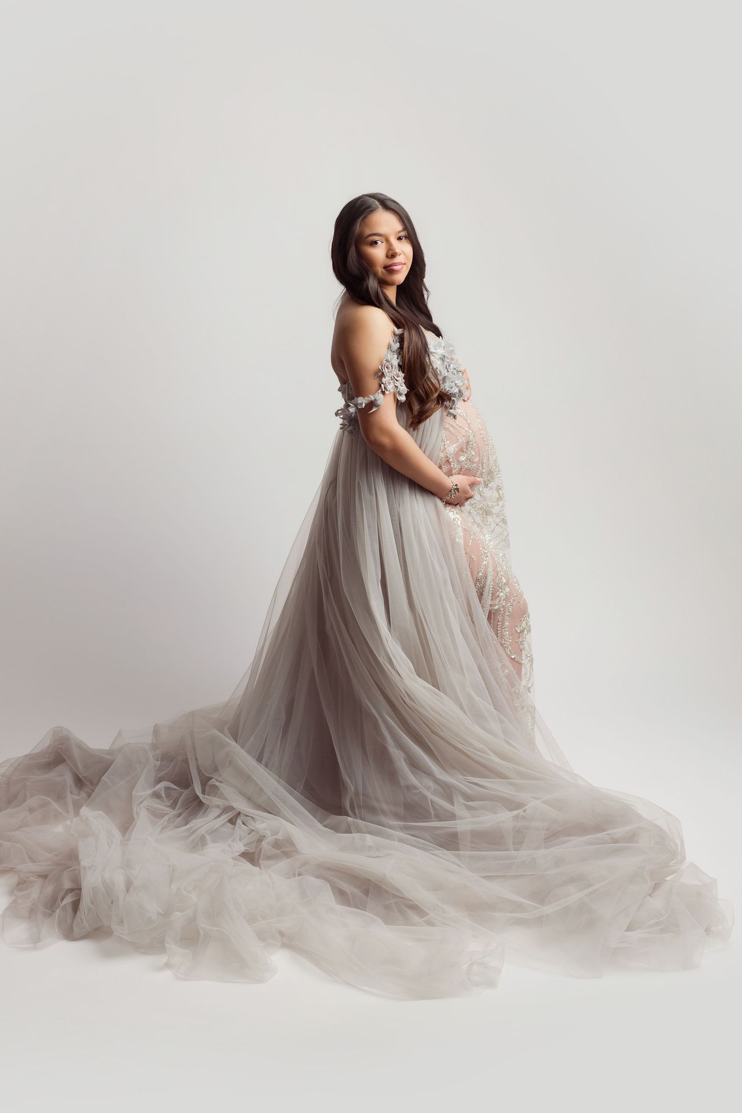 A figure in a flowing gray tulle gown poses gracefully against a white studio backdrop in an artistic portrait series.