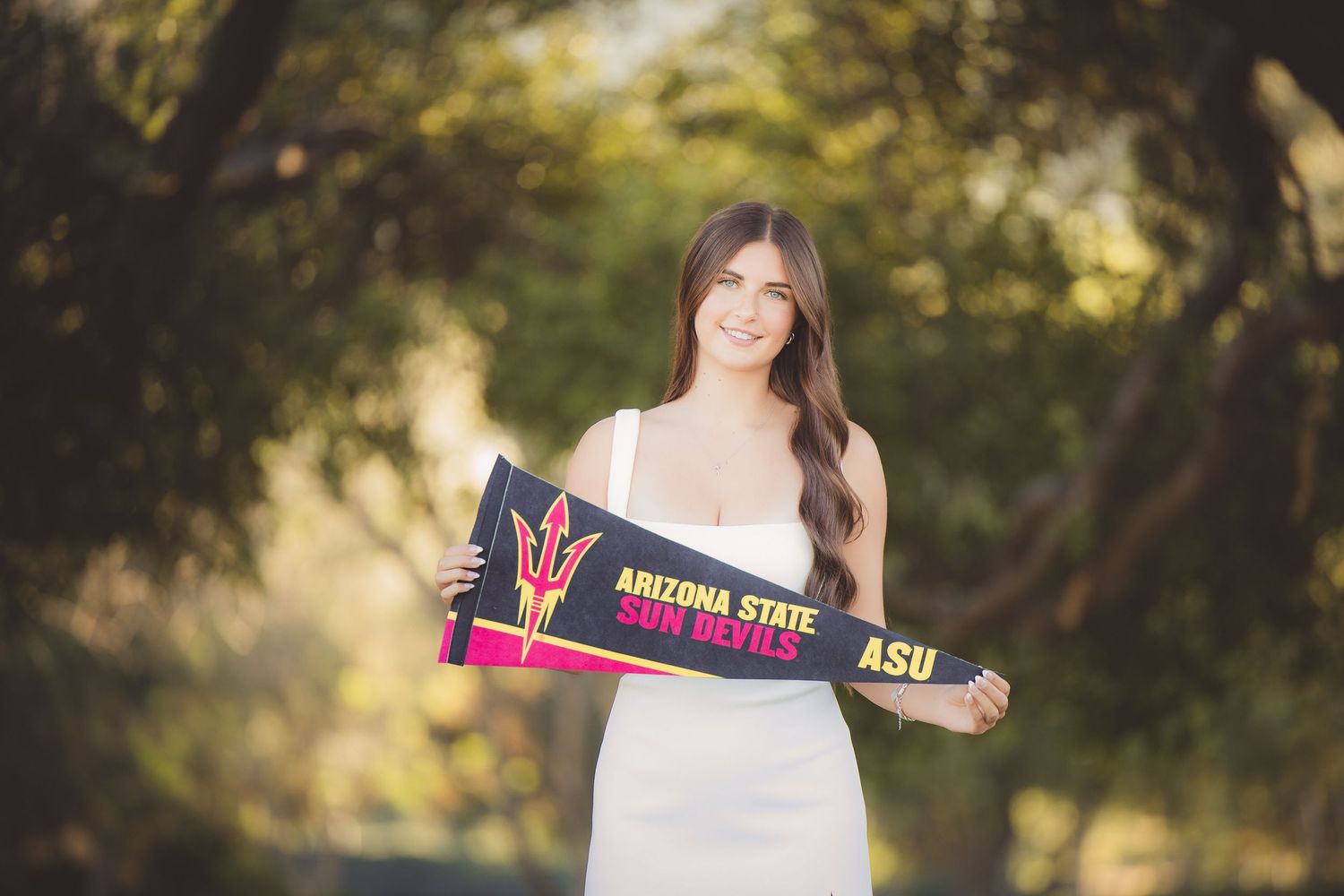Smiling young woman holds Arizona State Sun Devils pennant outdoors among green trees.