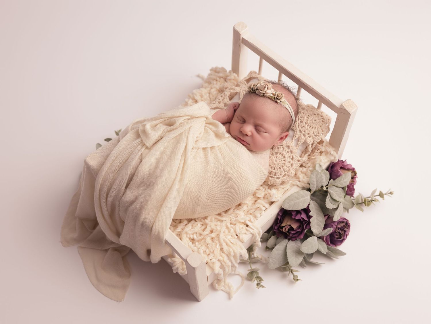 Newborn baby sleeps peacefully on miniature bed with cream blanket and purple floral accents during photo session.