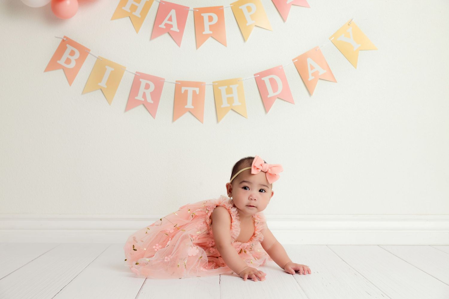 Baby girl in peach tutu dress crawling under a Happy Birthday banner during her first birthday cake smash photo session.