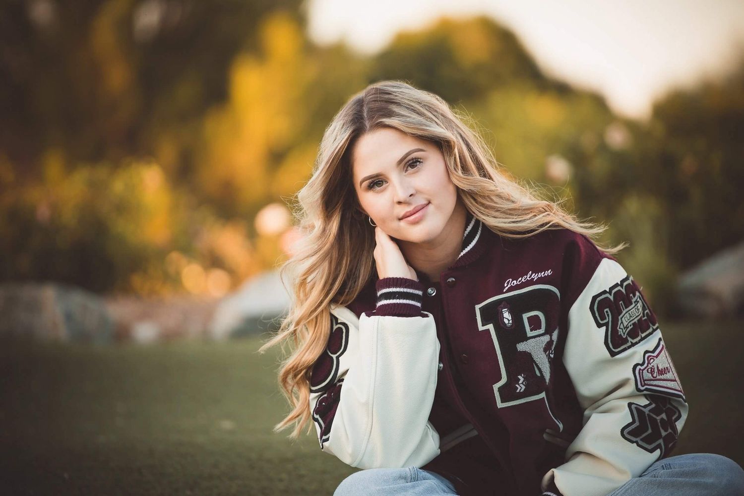 Young woman in varsity jacket sitting outdoors with bokeh background, smiling softly at camera.