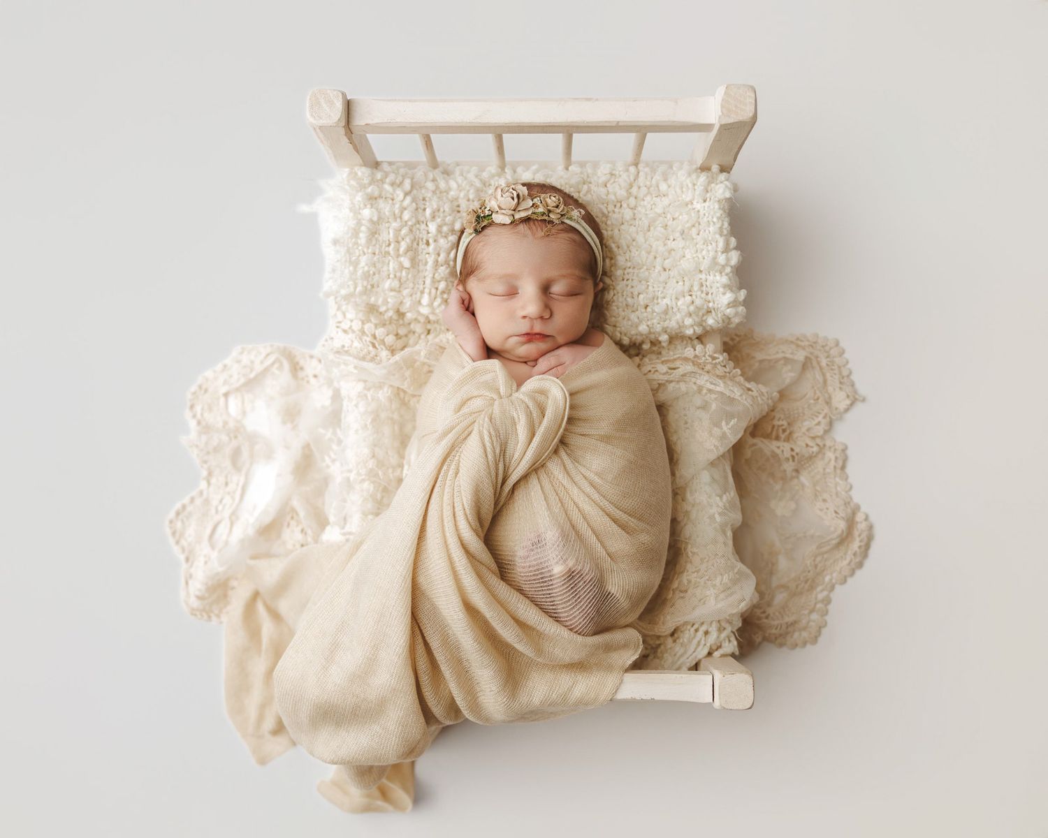 Newborn baby swaddled in cream wrap sleeping peacefully on a tiny white crib with lace pillow and floral headband.