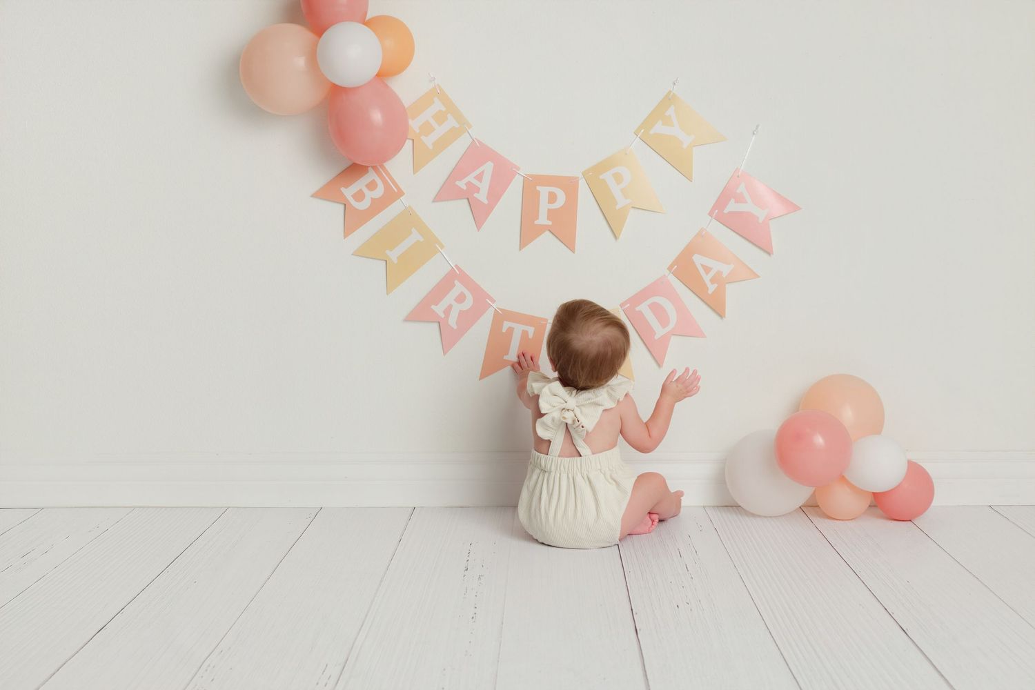 Adorable baby girl sitting on white floor reaching up to touch a pink Happy Birthday banner with balloons.