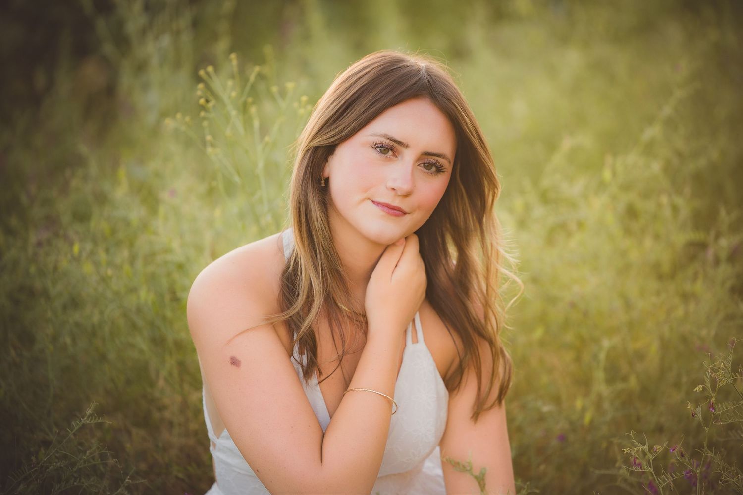 Young woman with long hair sitting in green meadow for outdoor senior portrait photography.