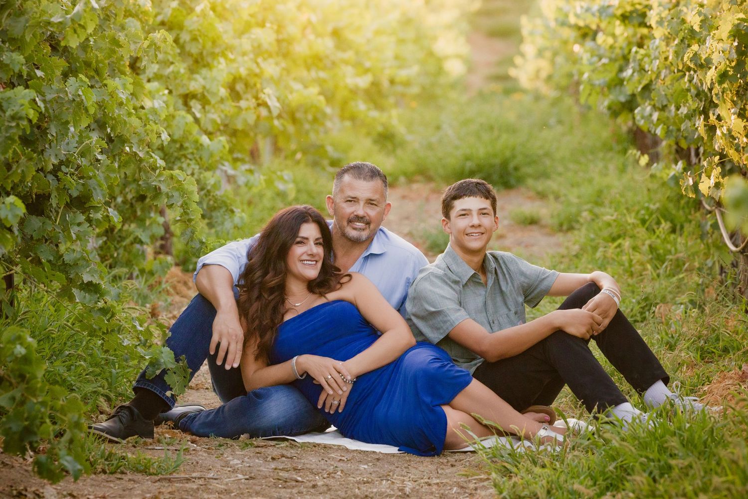 Three people sit together in a sunny vineyard with lush green vines on a warm summer evening.
