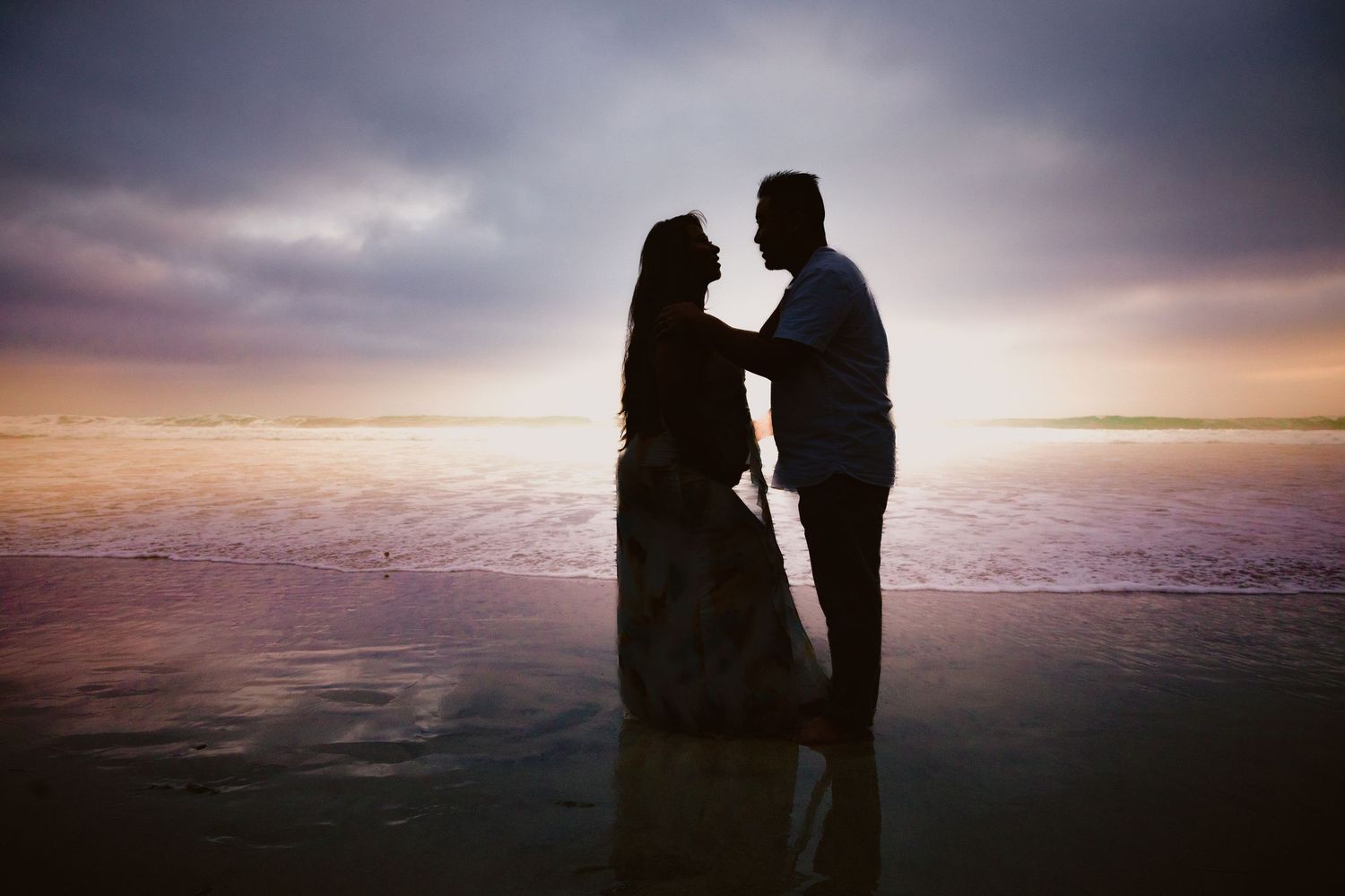 Silhouette of a romantic couple standing in shallow ocean water at sunset with dramatic purple sky.