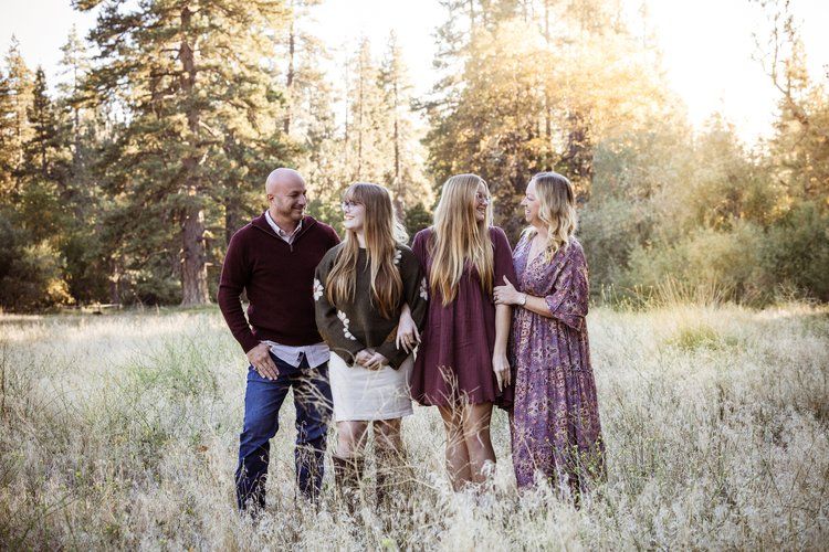 A family poses together in a sunlit meadow surrounded by pine trees wearing coordinating burgundy and neutral colors.
