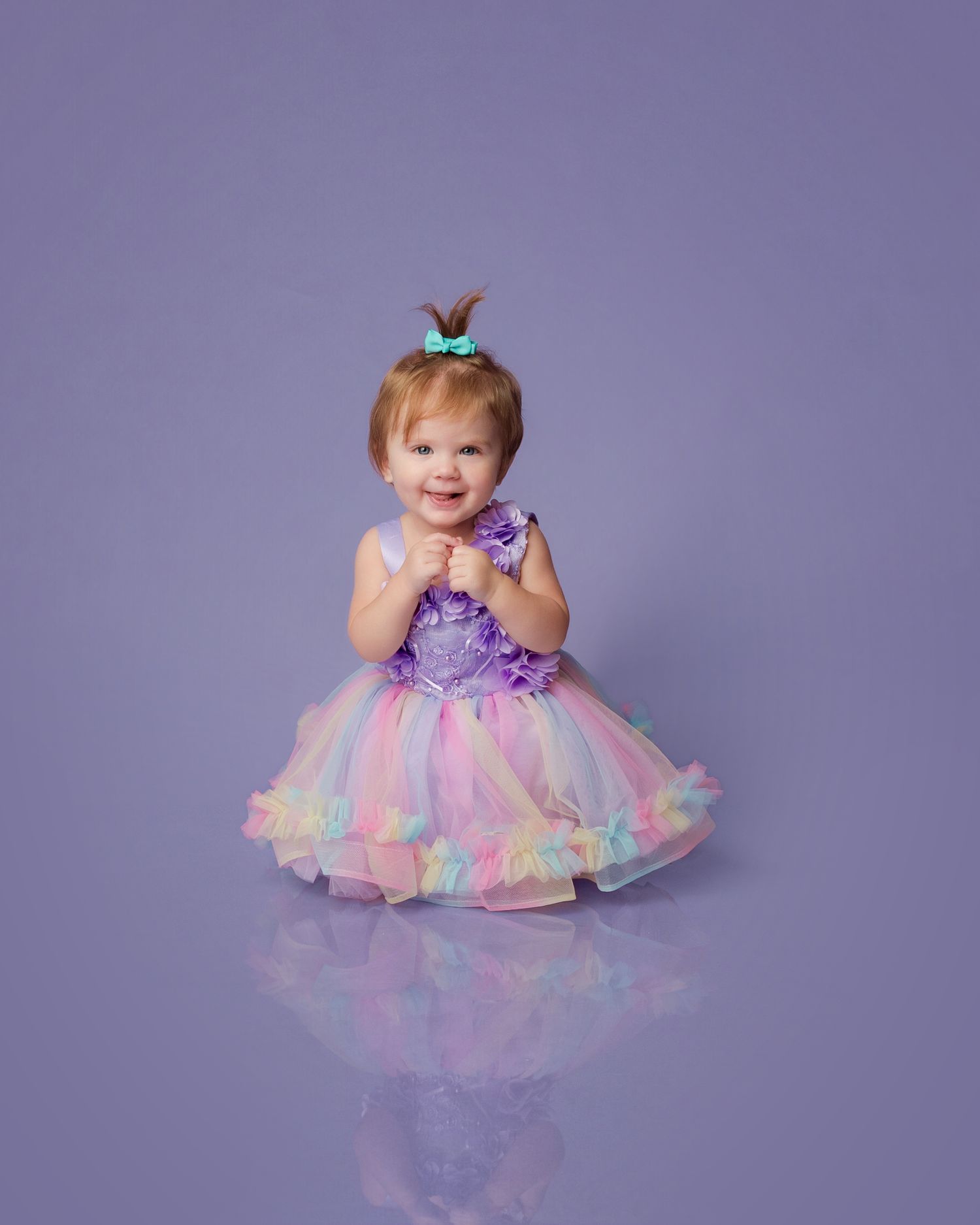 Smiling toddler girl in colorful rainbow tutu dress with purple bodice and teal bow on purple background.