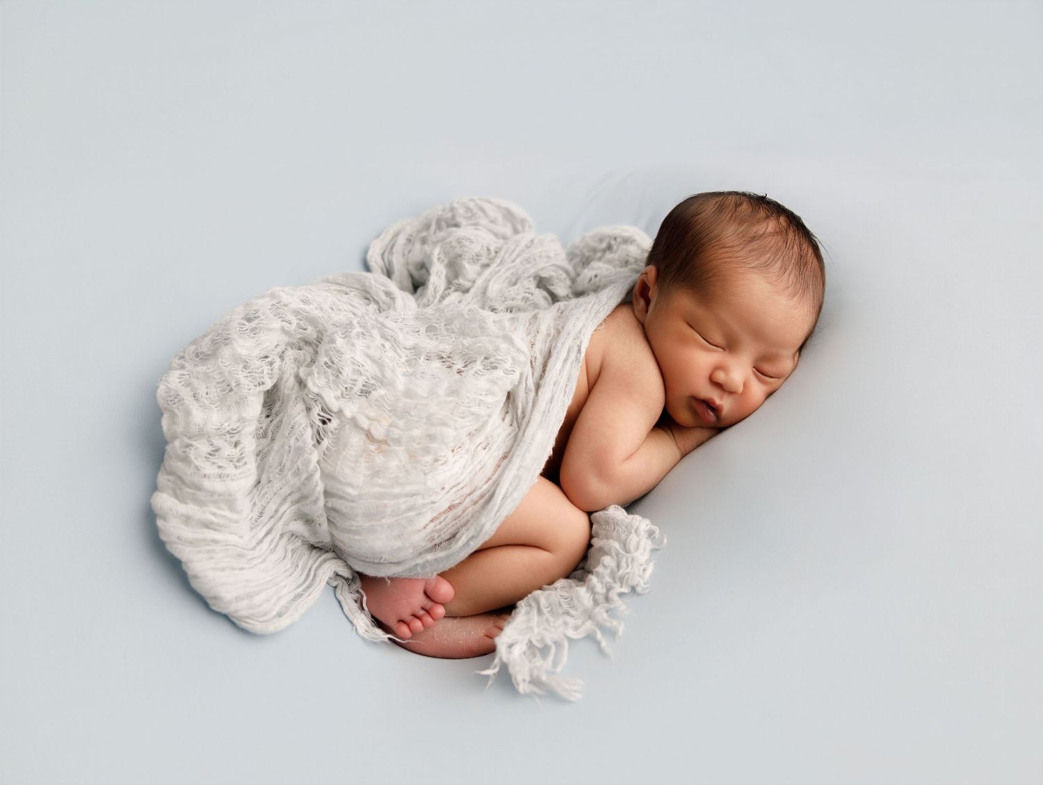 A sleeping newborn baby wearing white lace clothing lies peacefully on a soft white backdrop.