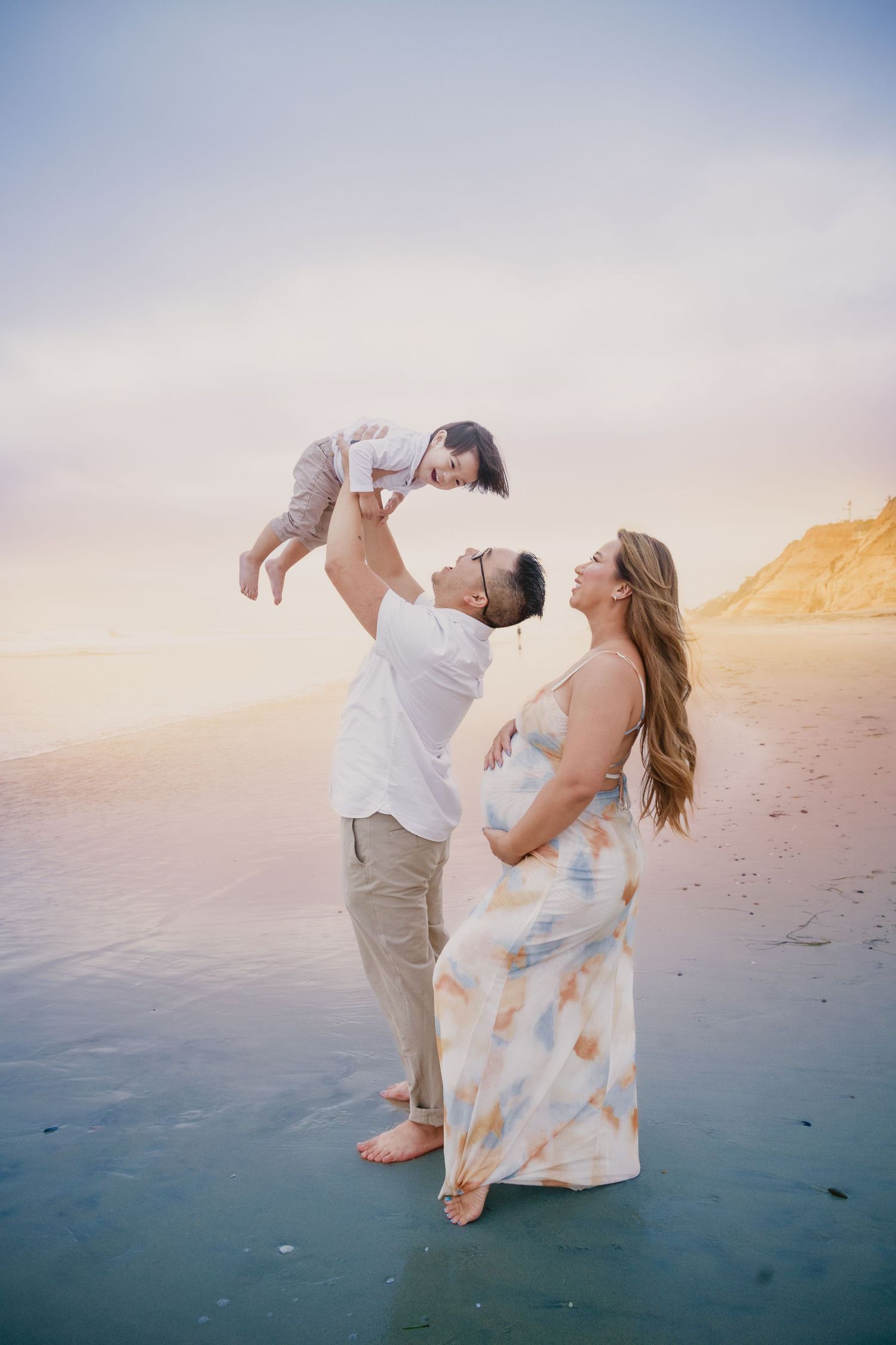 Pregnant woman in floral dress with partner lifting toddler on beach at sunset.