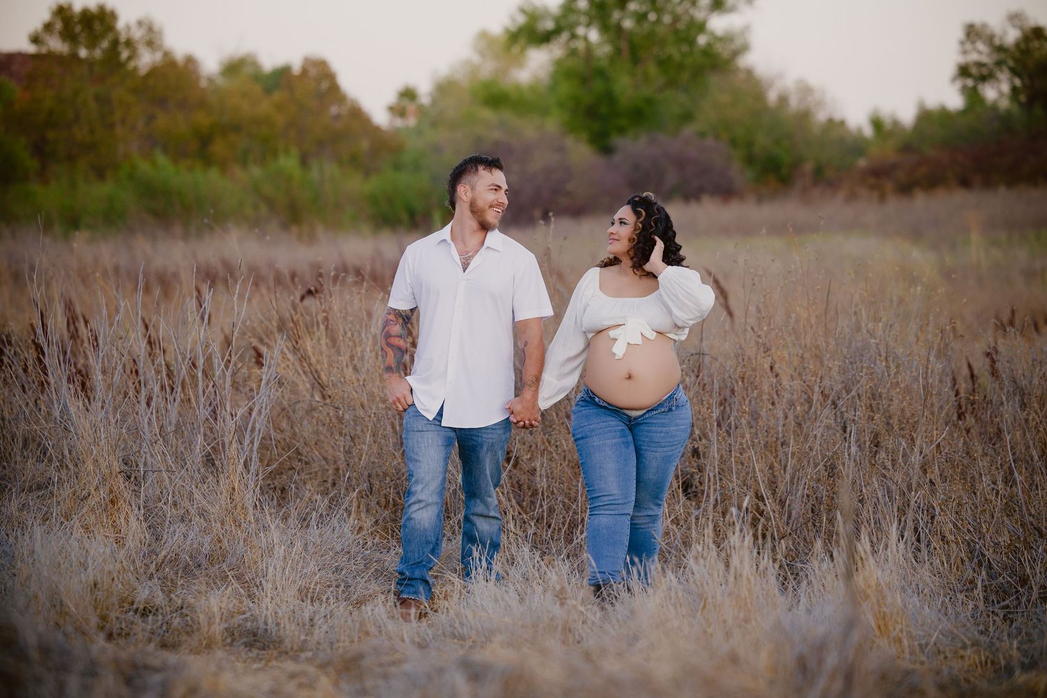 Expecting couple holds hands in golden field, woman showing baby bump, both wearing white shirts and jeans.