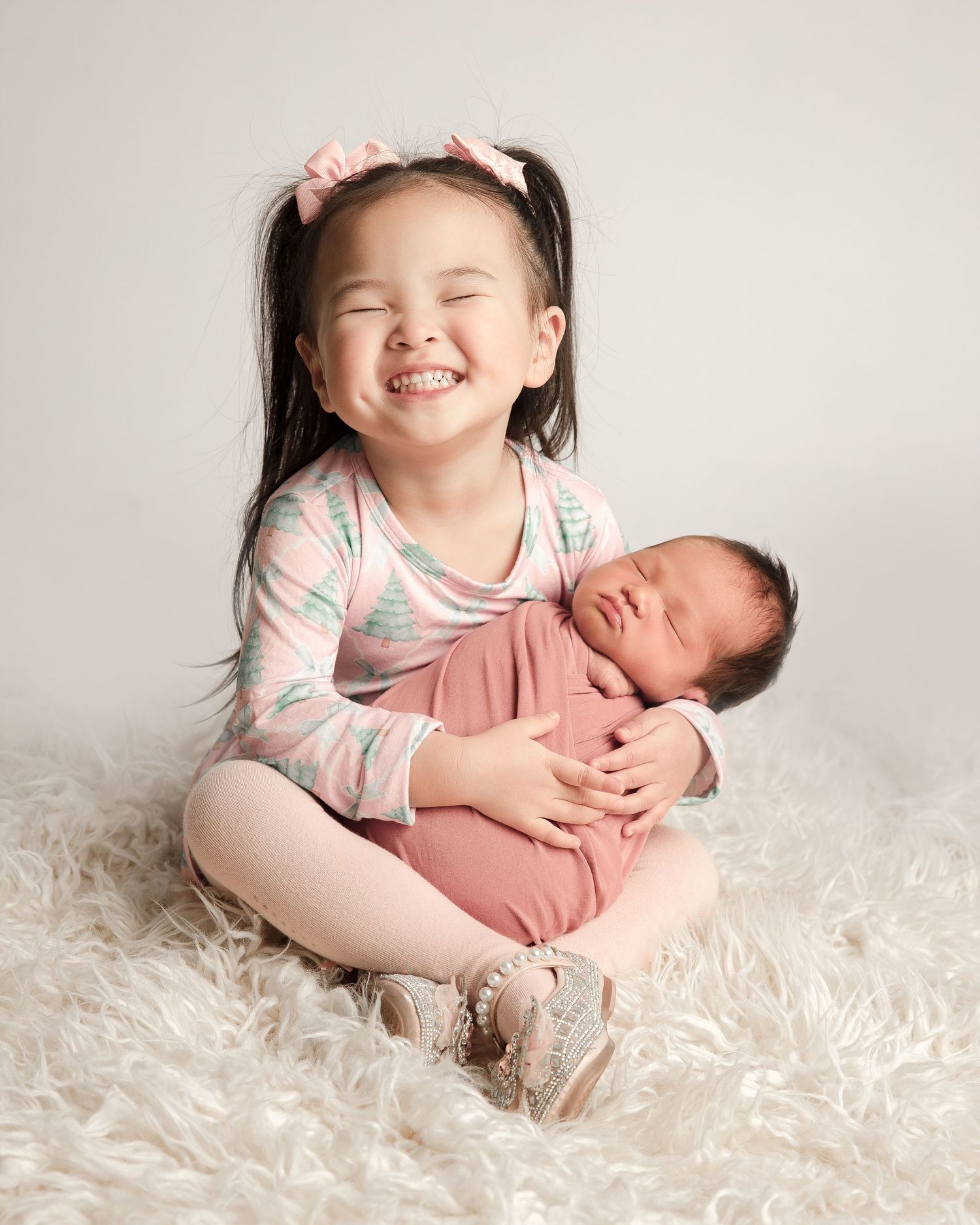 A young child in pink and white sitting on a fluffy rug while holding a swaddled newborn baby.