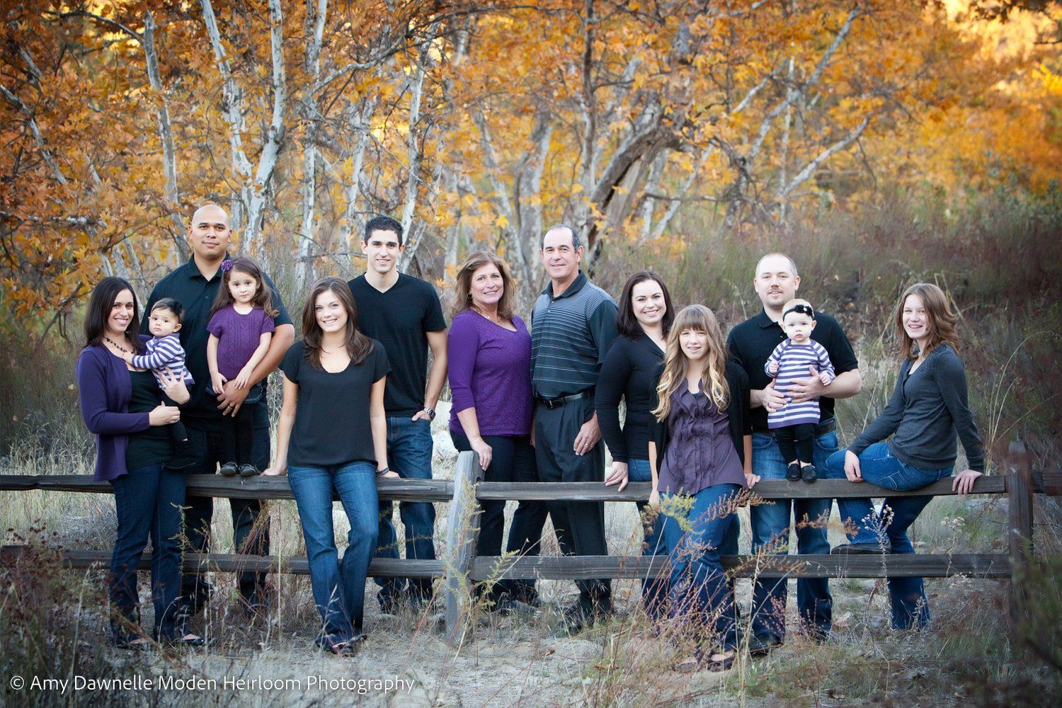 A large extended family poses together outdoors on a wooden fence with autumn birch trees and golden foliage in the background.