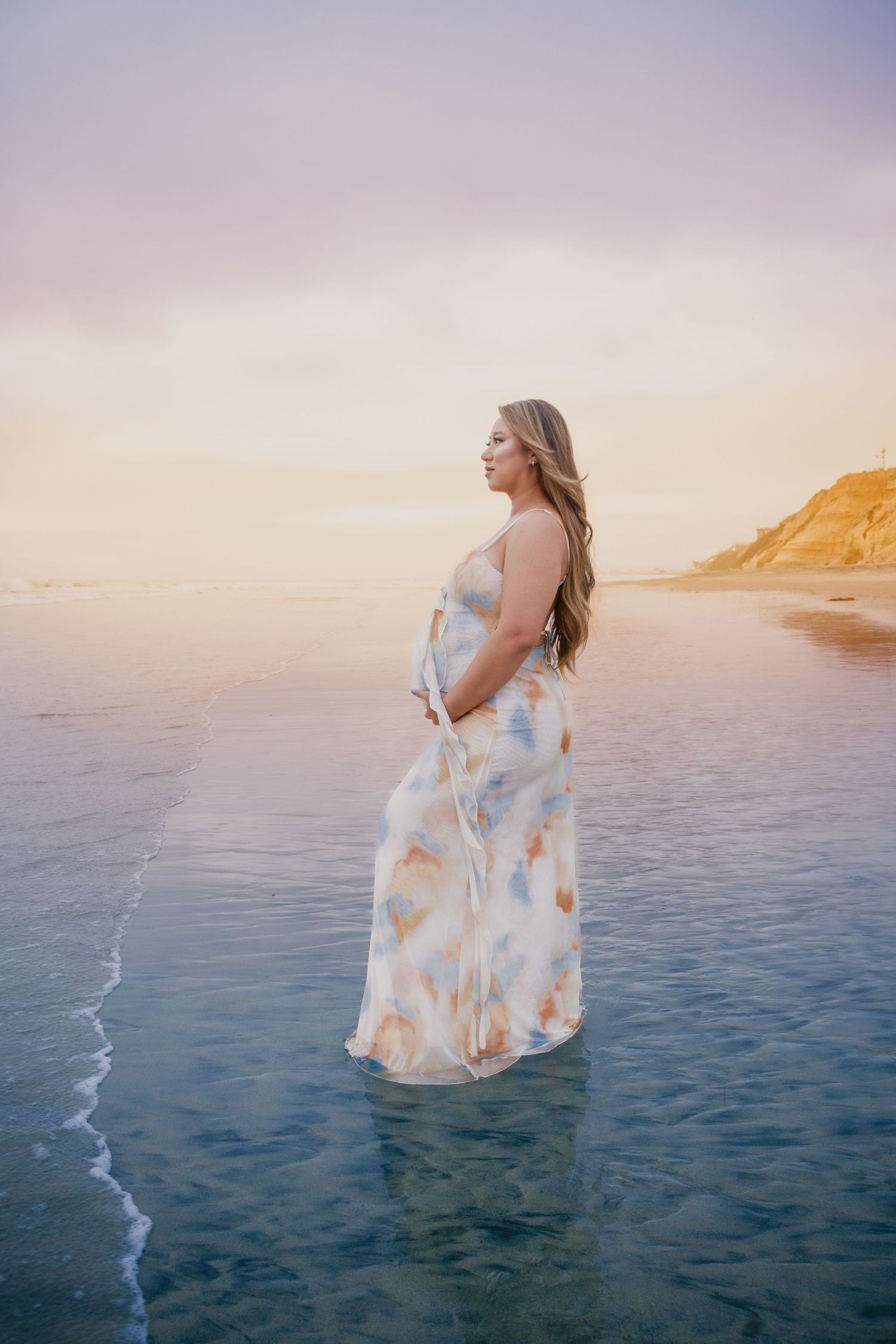 Pregnant woman in floral maxi dress standing in shallow ocean water at sunset on a beach.